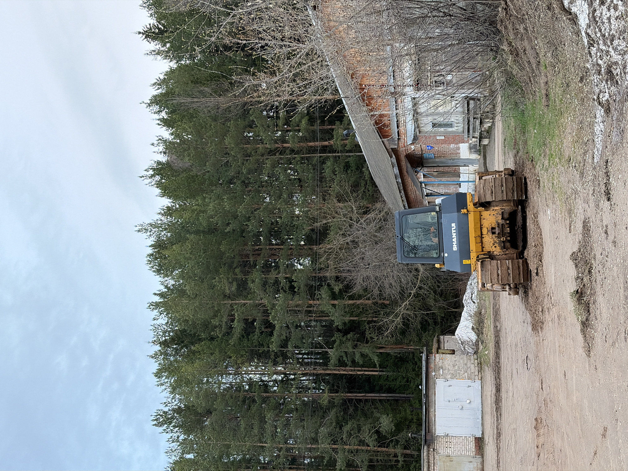 This image, taken in Chaikovsky, Russia, presents an outdoor scene under an overcast sky, suggesting late winter or early spring. A yellow and gray bulldozer, prominently displaying the brand "SHANTUI" on its cab, is parked on a dirt path in the right midground. Its heavy tracks are visible, caked with dirt.
To the left of the bulldozer, a dense forest of tall pine trees forms a significant natural backdrop, stretching across the left half of the image. Further into the background on the right, several older, utilitarian buildings, constructed from a mix of brick and concrete, are partially visible through a scattering of bare deciduous trees and shrubs. In the bottom right corner, a smaller concrete block building, possibly a garage or shed, features two light-colored metal doors.
The foreground consists of a wide, unpaved dirt road or area, showing tire marks and some muddy patches. Along the far right edge of this dirt area, there are remnants of patchy snow or ice mingled with dry grass and soil. The scene is static, with no human activity or movement visible.
