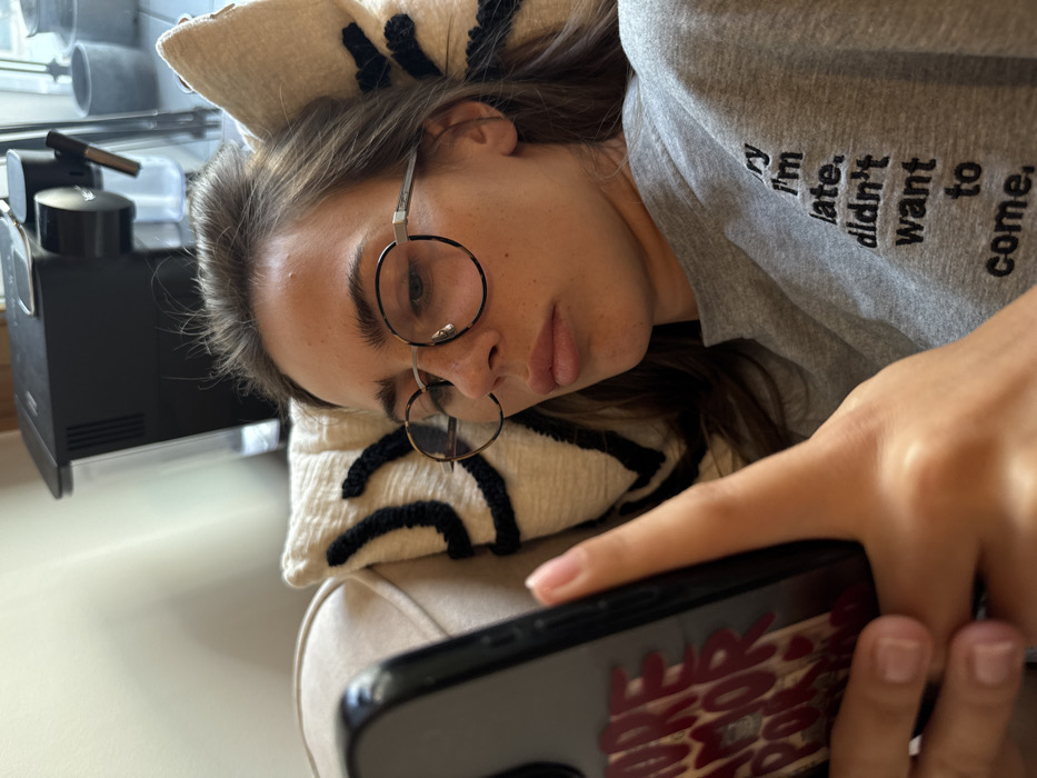 A female individual, estimated to be between 20 and 35 years old, is depicted reclining on a beige upholstered sofa in an indoor residential setting in Warsaw, Poland. Her head rests against a cream-colored throw pillow featuring a textured black abstract or geometric pattern. She wears a light grey, long-sleeved crew-neck sweatshirt with black embroidered text on the right chest area that reads: "sorry i'm late. didn't want to come." The subject has medium-brown hair and wears round eyeglasses with silver frames and tortoise-shell rims. Her expression is neutral, with slightly pursed lips, and her gaze is directed downward, focused intently on a black smartphone held in both hands. The phone's transparent case displays partially visible red text that reads "MORE AMOR POR F", suggesting "MORE AMOR POR FAVOR". Her right thumb is positioned over the phone's screen. In the background, to the subject's upper right, a black automatic coffee machine is visible on a surface next to a window or glass door. Beyond this transparent barrier, several dark-colored outdoor planters are partially visible. A plain light-colored wall occupies the upper left background. The scene suggests a relaxed, casual environment.