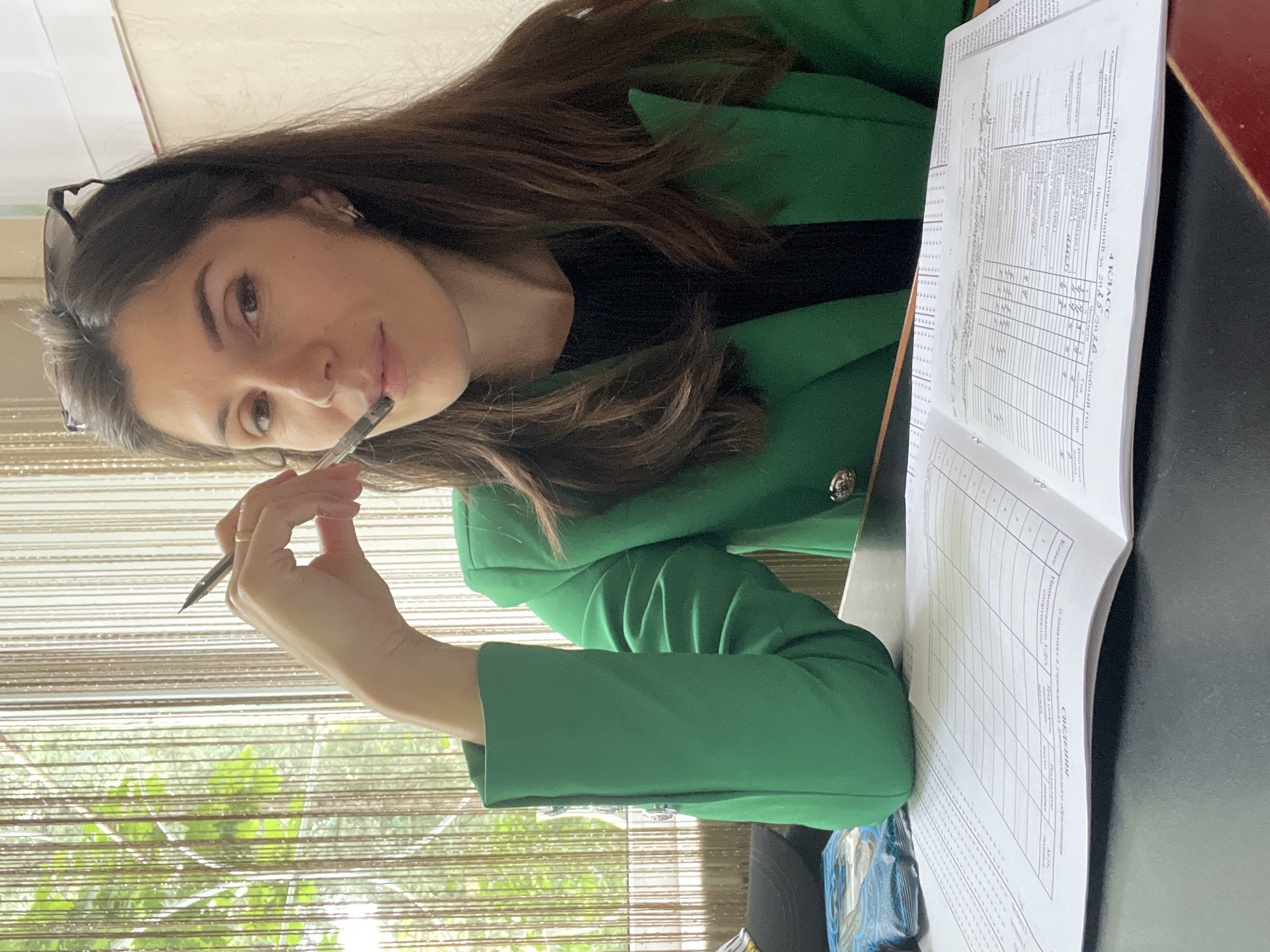 A young woman with long, dark brown hair and glasses resting on her head is captured in a close-up, thoughtfully posing with a pen held to her lips. She gazes directly at the viewer. She is wearing a black top under a vibrant green blazer with distinctive buttons on the cuffs. The scene appears to be an indoor setting in Kaspiysk, Russia, likely at a desk or table, with natural light streaming in from a window to her left, filtered by light-colored string or vertical blinds. Green foliage is visible through the blinds, suggesting it is daytime.
An open book, possibly a journal or a grade book, lies on a dark surface to her right. The pages feature a grid layout with Cyrillic text. Visible text on the book includes "4 КЛАСС" (4th Grade), "Табель оценки знаний за 2025 2026 учебный год" (Table of knowledge assessment for the 2025-2026 academic year), "Предметы" (Subjects), "Филолог" (Philologist), "Математик" (Mathematician), "Класс" (Class), and "СВЕДЕНИЯ" (INFORMATION), along with numbers like "3" and "119". The dates "2025 2026" indicate a contemporary or near-future context for the document. A small blue packet or box is also visible on the table. The overall impression is one of a person engaged in study or administrative work, pausing for a reflective moment.