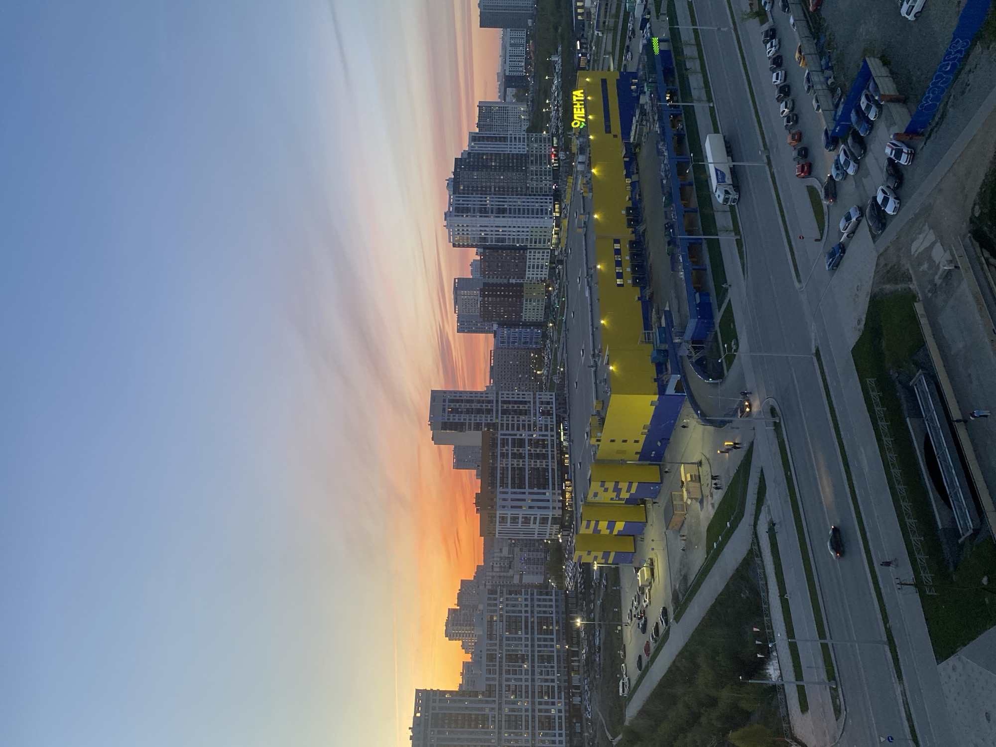 The image shows an aerial view of a city at dusk. The sky transitions from a blue to orange gradient. Buildings, mostly residential high-rises, line the horizon. A large, yellow and blue building is centrally located in the foreground, identified by the Cyrillic text "ЛЕНТА" (Lenta) on its facade. Adjacent to the Lenta building is a large parking lot containing many vehicles. Streets with light traffic run beside the building and parking areas. The location is Yekaterinburg, Russia, identified by the ЛЕНТА (Lenta) superstore.