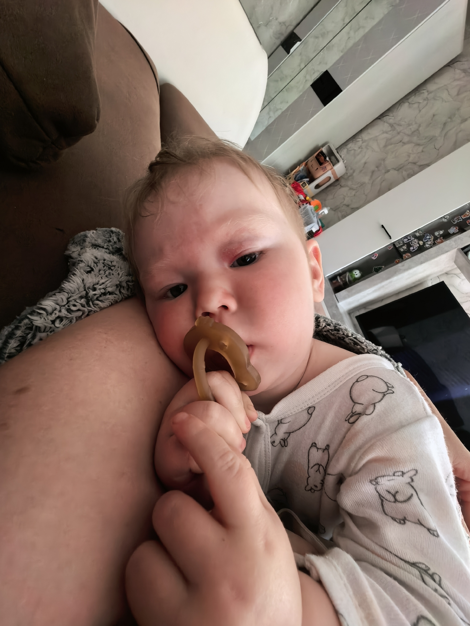 A baby with fair skin and light hair is lying on a person's lap, holding a brown pacifier in their hand. The baby is wearing a white onesie with a pattern of sketched dogs. The person's lap is covered with a patterned blanket. In the background is a marble-like wall, white cabinets, and a dark rectangular shape, possibly a TV screen. A toy truck and other small objects are on a shelf. The image was taken in an indoor setting. The location, Sterlitamak, Russia, is not discernible from the image.