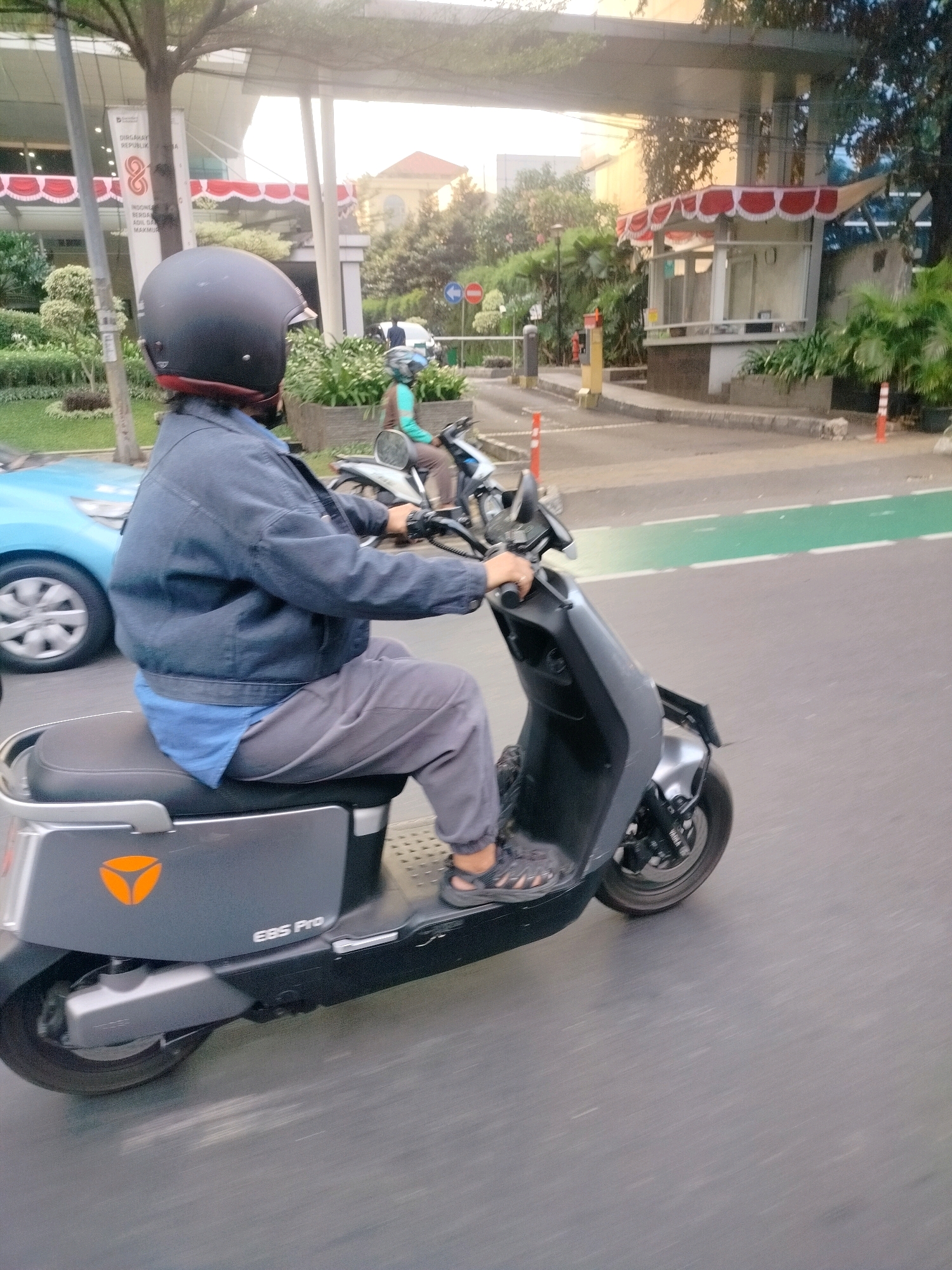 A person wearing a black helmet and a dark blue jacket rides a grey electric scooter. The scooter has an orange logo on its side and the text "EBS Pro". The person is wearing grey pants and sandals. The scooter is on a paved road with a green lane marker. In the background, a blue car is visible, as is a building with red and white awnings. The scene appears to be in a city, possibly East Jakarta, Indonesia, given the context of the surrounding environment and vehicle types.