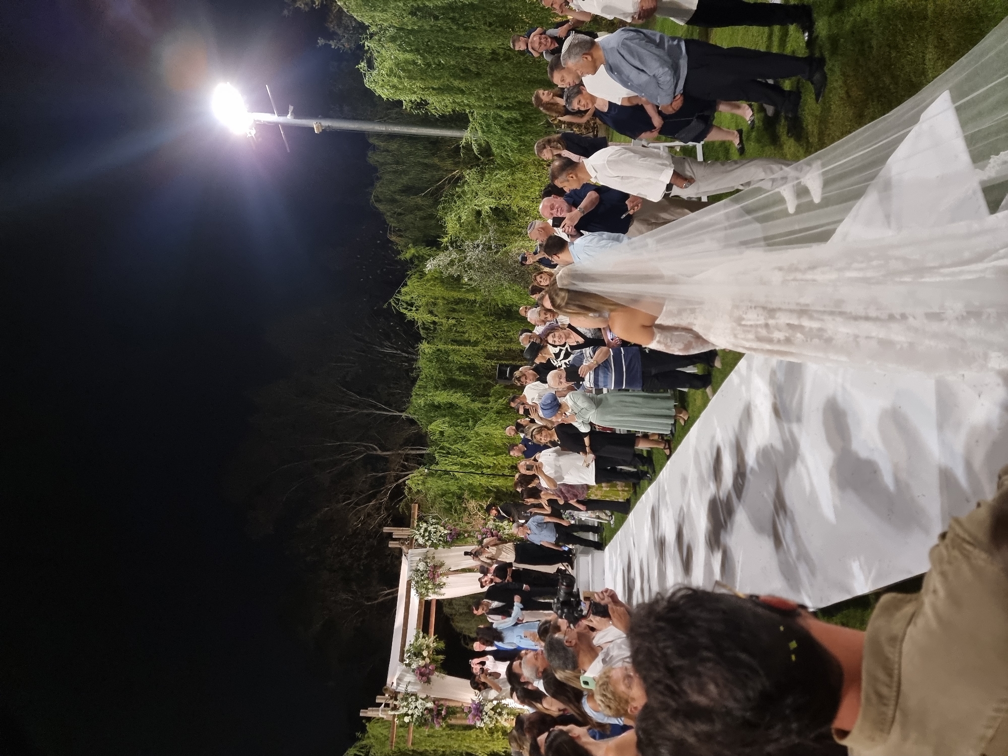 The image depicts an outdoor wedding ceremony at night in Ga'ash, Israel. A white carpet stretches down a grassy aisle, lined by guests observing the proceedings. A floral arch marks the end of the aisle where a bride, in a white lace wedding gown and veil, is walking. She is accompanied by a man in a suit. Other individuals, including a photographer with a professional camera, are positioned near the arch. Guests are scattered on both sides of the aisle, many holding up their phones to capture the moment. The background is filled with lush green trees and the illumination from a tall street lamp suggests the event is taking place after sunset.