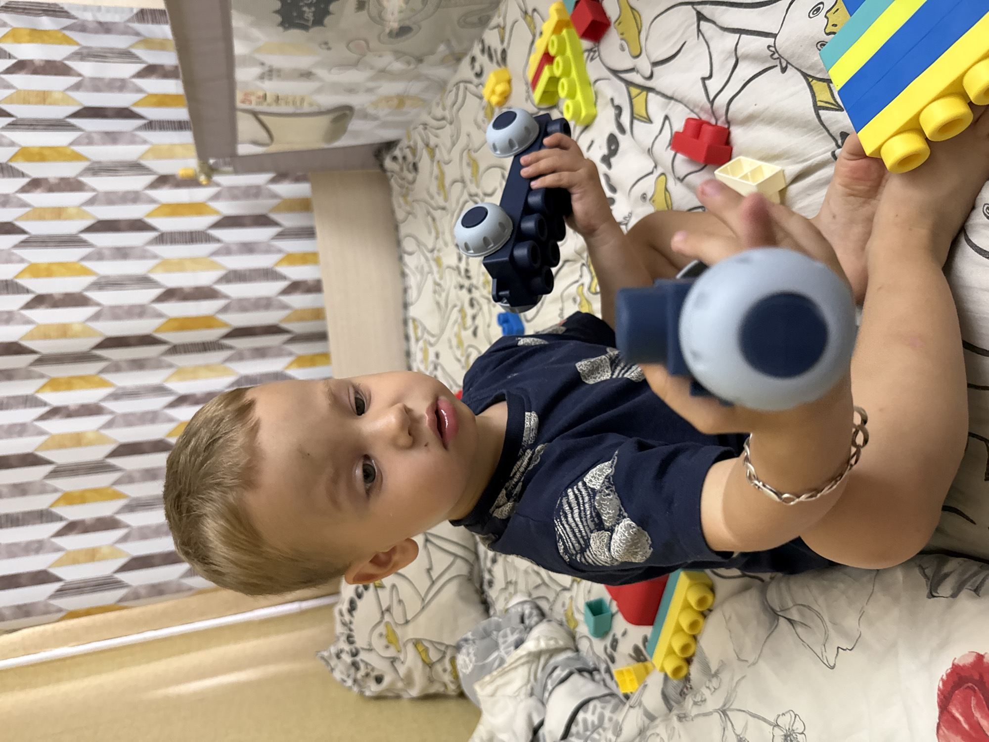 A young boy with blond hair and blue eyes is sitting on a bed, holding a dark blue toy car with two white wheels and black cylindrical details. He is wearing a dark blue t-shirt with a gray pattern. The bed has white sheets with a gray and white duck pattern. Scattered around him are colorful building blocks, including yellow, blue, and red pieces. The background shows a wall with a patterned curtain. The boy is looking down at the toy he is holding, with his mouth slightly open. A silver bracelet is visible on his left wrist. The scene appears to be indoors, likely in a bedroom. The location is identified as Rusanovo, Russia.