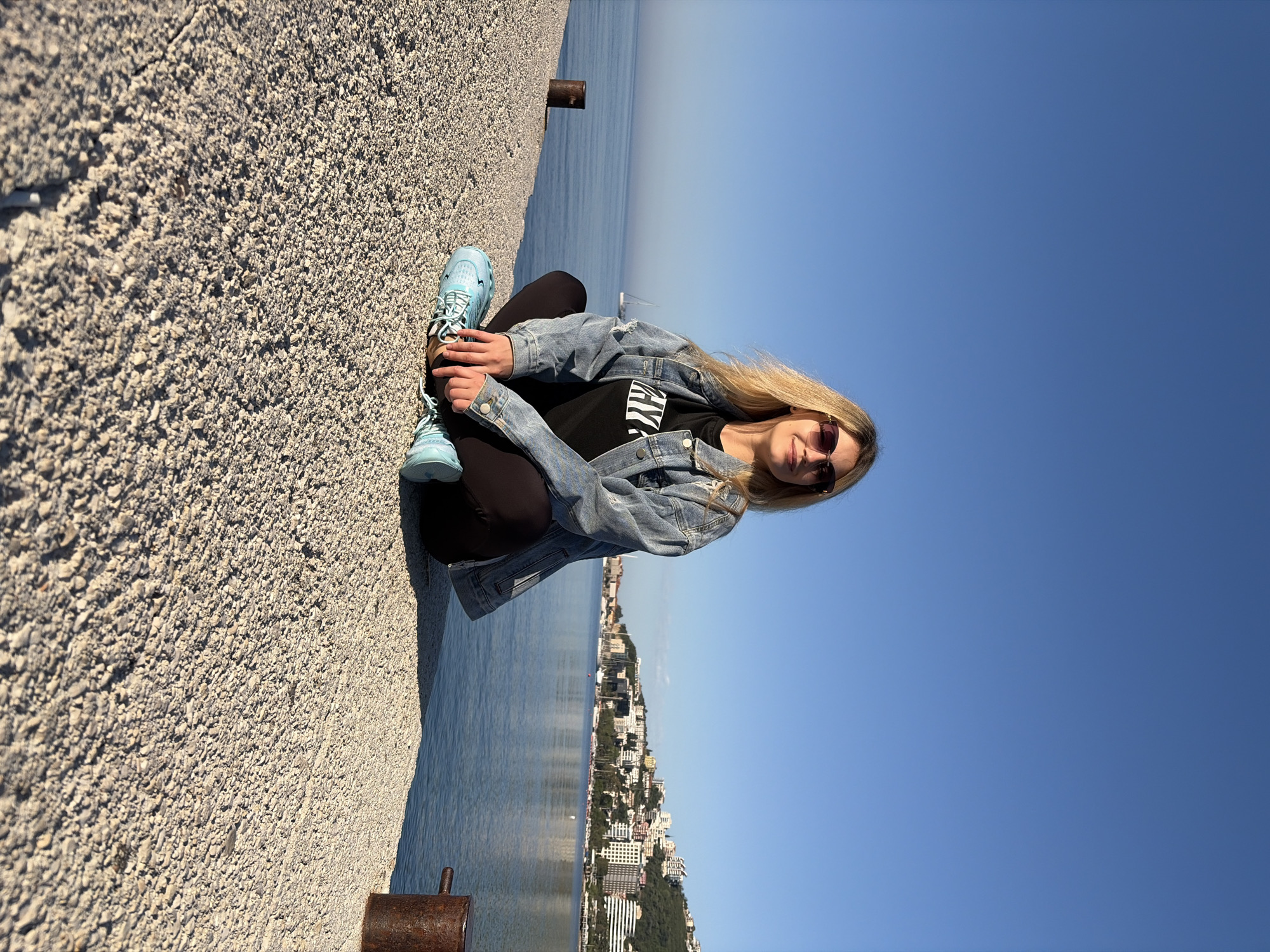 A woman sits with her legs crossed on a concrete wall overlooking the Adriatic Sea and the city of Budva, Montenegro. The wall is made of small, light-colored aggregate concrete. She wears black leggings, a black t-shirt with white lines, a light blue denim jacket, light blue sneakers, and sunglasses. The background is the blue sky, the sea, and a city with white buildings along the coastline. A metal mooring bollard is visible on the wall near the woman's feet. The angle of the image is tilted.