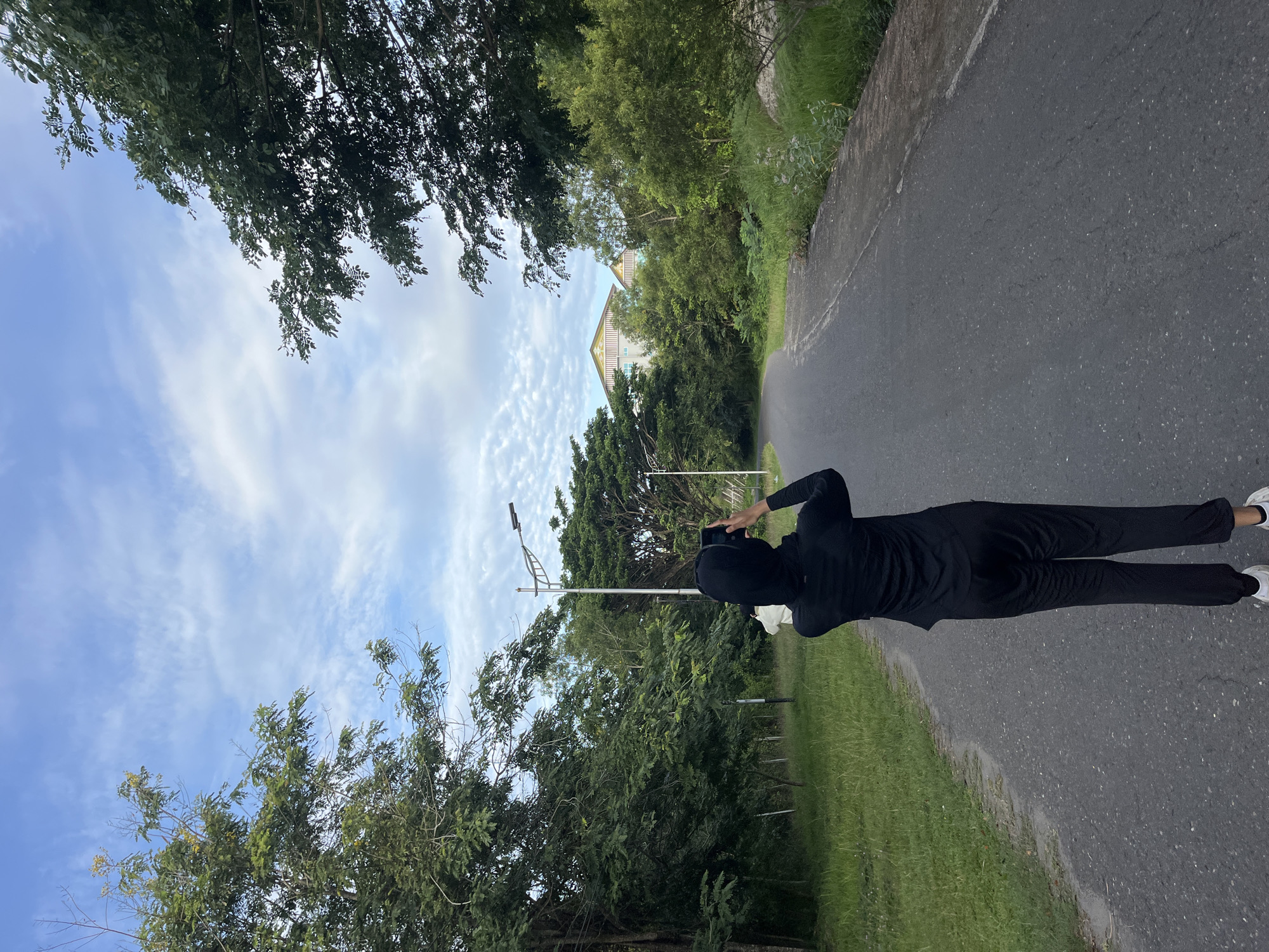 A person wearing a black hooded top and pants stands on the asphalt road. The person is holding a smartphone, presumably taking a photo or video. The person is located on a road surrounded by trees and greenery. Overhead power lines and poles are located near the road. In the distance, the roof of a building is visible. The sky is blue and cloudy. Based on the location information given, this scene may be somewhere in Lhokseumawe, Indonesia.