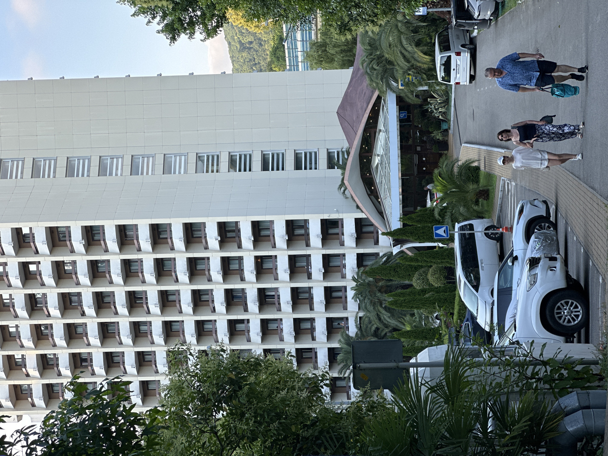 The image depicts a scene in Khostinskiy, Russia, featuring a tall, modern hotel building with numerous balconies. In the foreground, a paved road leads towards the hotel, with several white cars parked along the left side. A man, a woman, and a younger person are walking away from the camera on the right side of the road. The man, wearing a blue patterned short-sleeved shirt and black shorts, carries a blue bag. The woman, in a black tank top and patterned trousers, walks beside him. The younger person, wearing a white cap, white t-shirt, and white shorts, walks slightly ahead of the woman. Lush green trees and palm trees are interspersed throughout the scene, with a distant hillside visible in the background. A prominent blue pedestrian crossing sign is visible on the right side of the road.