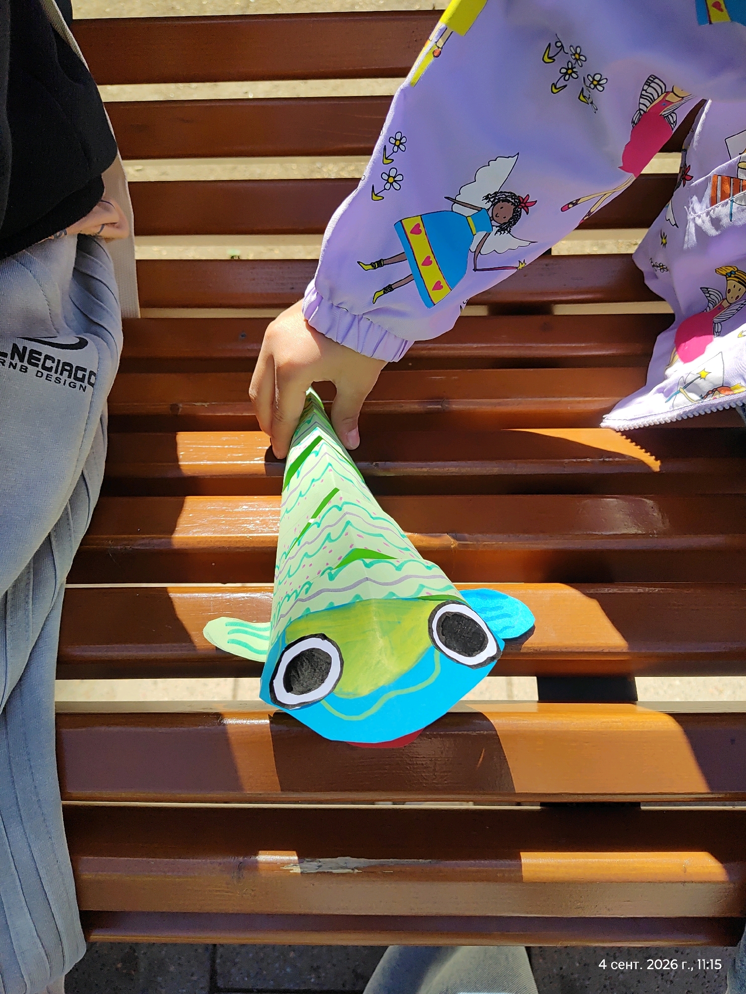 Two individuals are seated on a brown wooden park bench. One individual, wearing a light grey sweatpant with the logo "NECIAGO RN B DESIGN," is partially visible on the left side of the frame. The other individual, wearing a light purple jacket adorned with cartoon fairy designs, is on the right. They are holding a colorful, handmade paper fish decoration. The fish is cone-shaped, predominantly light green with blue accents, and features two large black and white eyes. The bench is situated outdoors, with sunlight casting shadows across its surface. The background suggests an urban park setting. The timestamp "4 сент. 2026 г., 11:15" is overlaid in the bottom right corner, indicating the date and time. The location of Astana, Kazakhstan, is not visually discernible from the image.