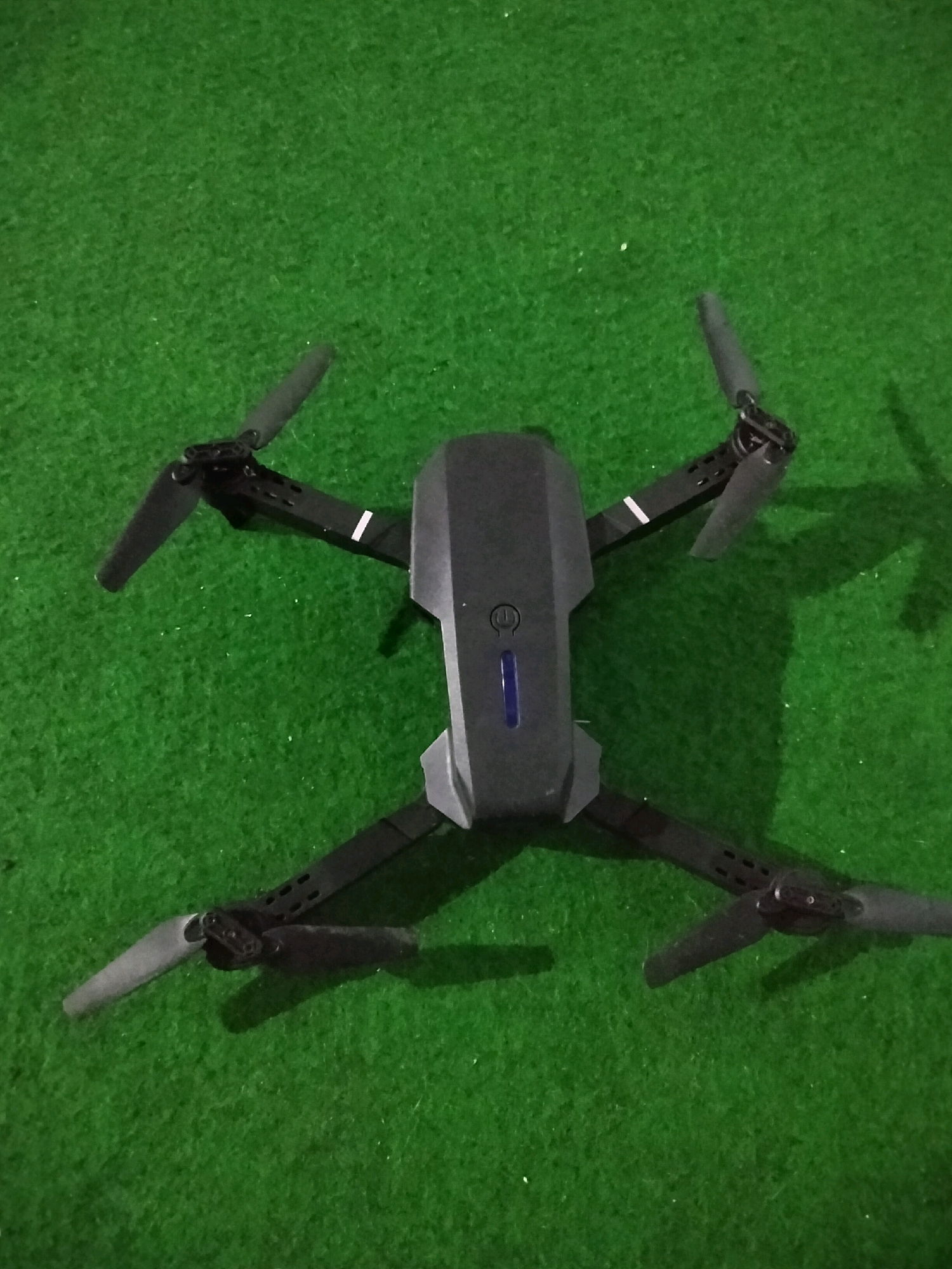 A drone is positioned on a green, textured surface that appears to be artificial grass. The drone is dark gray and has four arms, each with a black propeller. The body of the drone is centrally located, with a blue light visible on its surface. The drone's arms are extended outwards, forming an 'X' shape. Shadows of the drone are cast on the green surface. The image does not contain any discernible landmarks or environmental features that would indicate a specific location within Prapag Kidul, Indonesia. The scene is static and focuses solely on the drone.