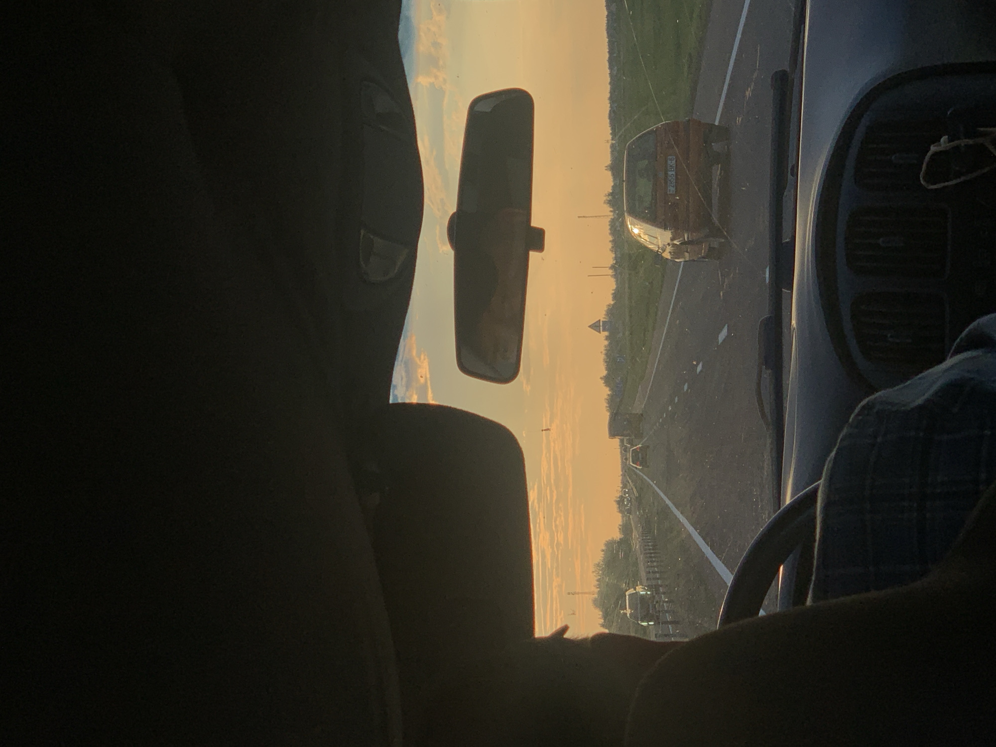 The image captures a view from inside a vehicle traveling on a highway during sunset. The perspective is from the driver's seat, with a portion of the steering wheel and dashboard visible in the foreground. A person's shoulder and head are partially visible on the left side, suggesting the driver or a passenger is in the frame. In the middle ground, other vehicles are seen on the road, including a reddish-orange car in the right lane. The sky is illuminated by the setting sun, casting a warm, orange glow across the scene and creating a hazy atmosphere. The landscape consists of flat terrain with some vegetation and structures, including what appears to be a small building or tower in the distance on the left. The overall impression is of a journey on a rural highway in Belarus at dusk.