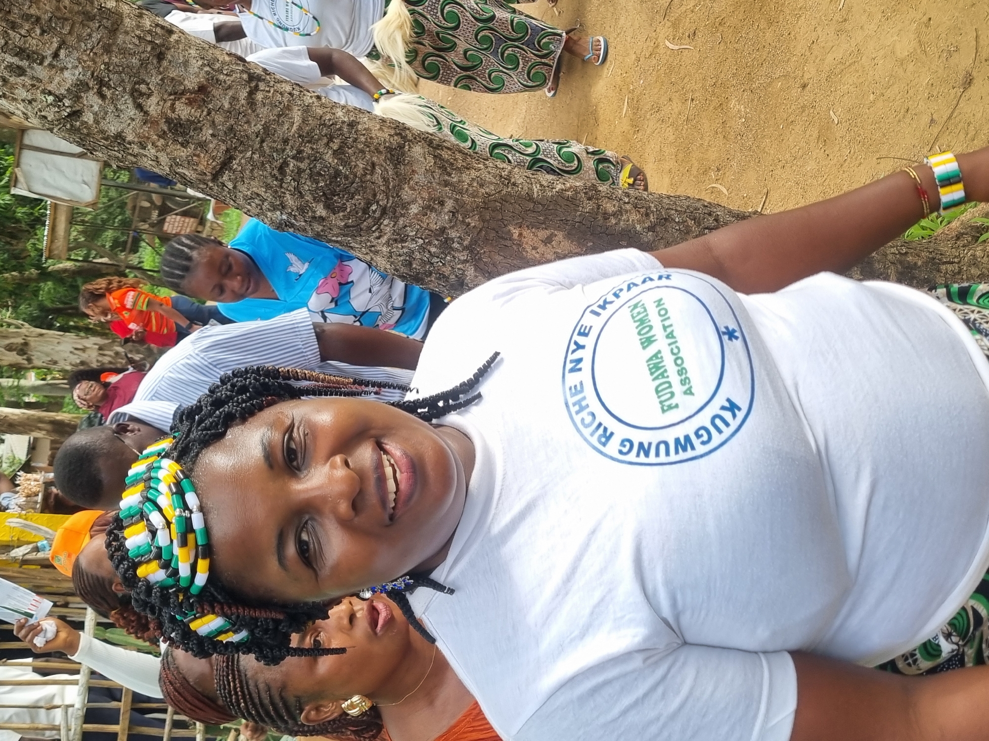 The image shows a group of people outdoors, possibly at a gathering or event. In the foreground, a woman with dark skin and braided hair adorned with colorful beads smiles at the camera. She is wearing a white t-shirt with a circular logo that reads "KUGMUNGM RICHE NYE IKPAAR FUDAMA WOMEN ASSOCIATION". Behind her, other individuals are visible, some with their faces partially obscured. The environment appears to be dusty with trees and foliage in the background, suggesting an outdoor setting in a city like Jos, Nigeria. The ground is a sandy, earthen color. The overall impression is one of a community event.