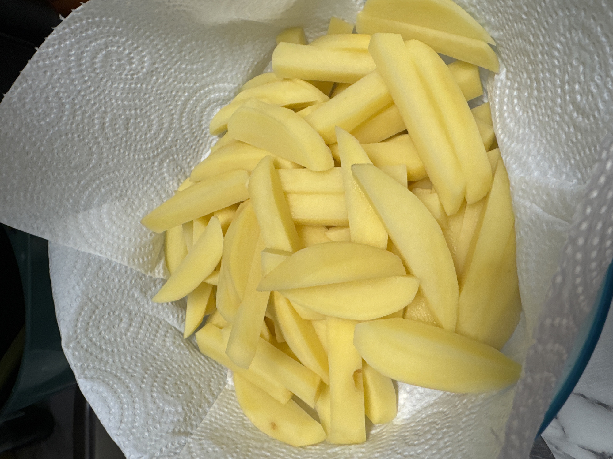 A close-up, overhead shot captures a pile of thinly sliced, uncooked potatoes resting on a white paper towel. The potatoes are bright yellow and appear to have been cut into uniform, baton-like shapes. Some of the potato slices exhibit a slightly curved or wedge-like appearance. The paper towel is textured with a circular pattern of embossed dots. In the background, a dark, blurred object, possibly a kitchen counter or appliance, is visible. The lighting is bright, highlighting the fresh cut of the potatoes.