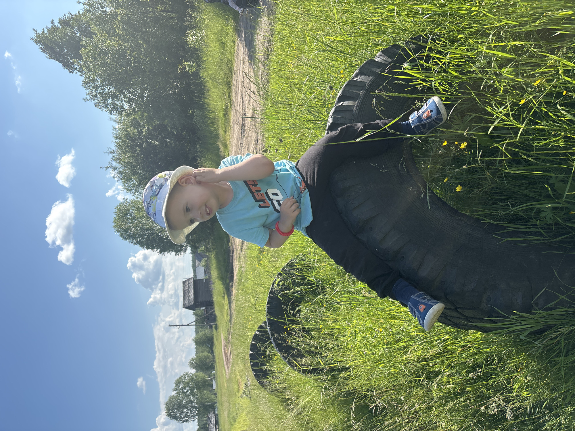 A young boy wearing a blue t-shirt, black pants, and a patterned bucket hat sits on a large tire in a grassy field. He is facing to the left, with his right hand touching his ear. His left leg is bent, and his blue shoes are visible. Two other large tires are partially submerged in the tall grass behind him, and a dirt road curves in the background. Trees and a small wooden structure are visible in the distance. The sky is blue with scattered white clouds. This scene is likely located in the rural outskirts of Kurgan, Russia, given the wooden buildings and natural landscape.