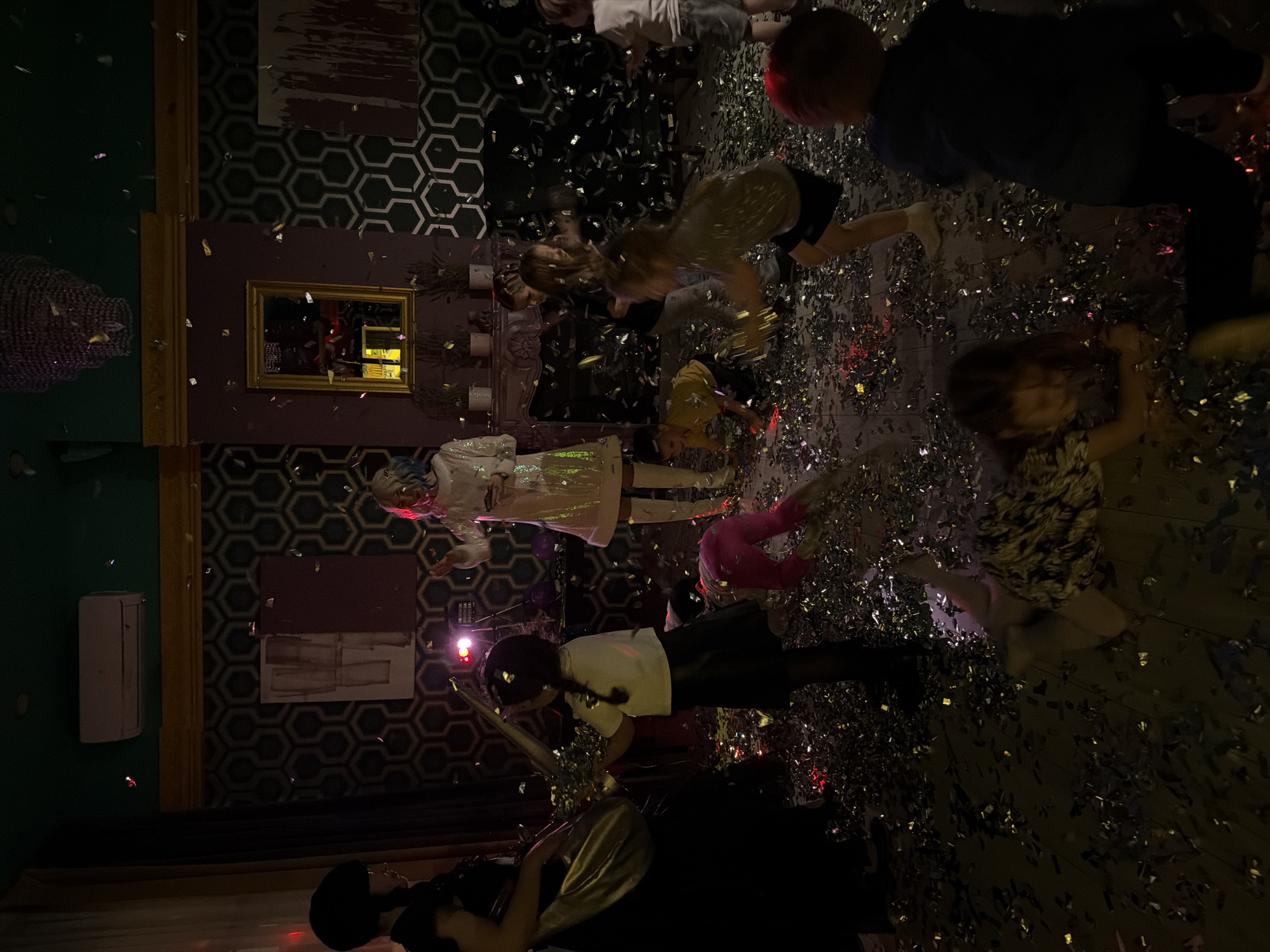 An indoor scene in Khimki, Russia, depicts an event primarily involving children, within a dimly lit room. The wooden floor is extensively covered with metallic confetti, and additional confetti is visible in mid-air. Approximately 10-12 individuals are present, consisting of at least two adults and several children. On the left, an adult female with dark hair wears a dark garment and carries a large, metallic, reflective object. In the central background, another adult female with light-colored hair is dressed in a white, sequined garment and white boots; she is positioned near a bright light source, possibly a projector, and a speaker unit. Children of varying young ages are dispersed throughout the confetti-covered floor, engaging with the confetti; one child in a white shirt and dark skirt operates a confetti cannon. Other children are observed crawling or standing. The room's interior features dark green hexagonal wallpaper on the upper walls, a dark plum lower wall section delineated by a gold trim, and a prominent golden-framed mirror situated above a mantelpiece adorned with small plants. A large crystal chandelier hangs from the dark green ceiling, where a white air conditioning unit is also mounted.