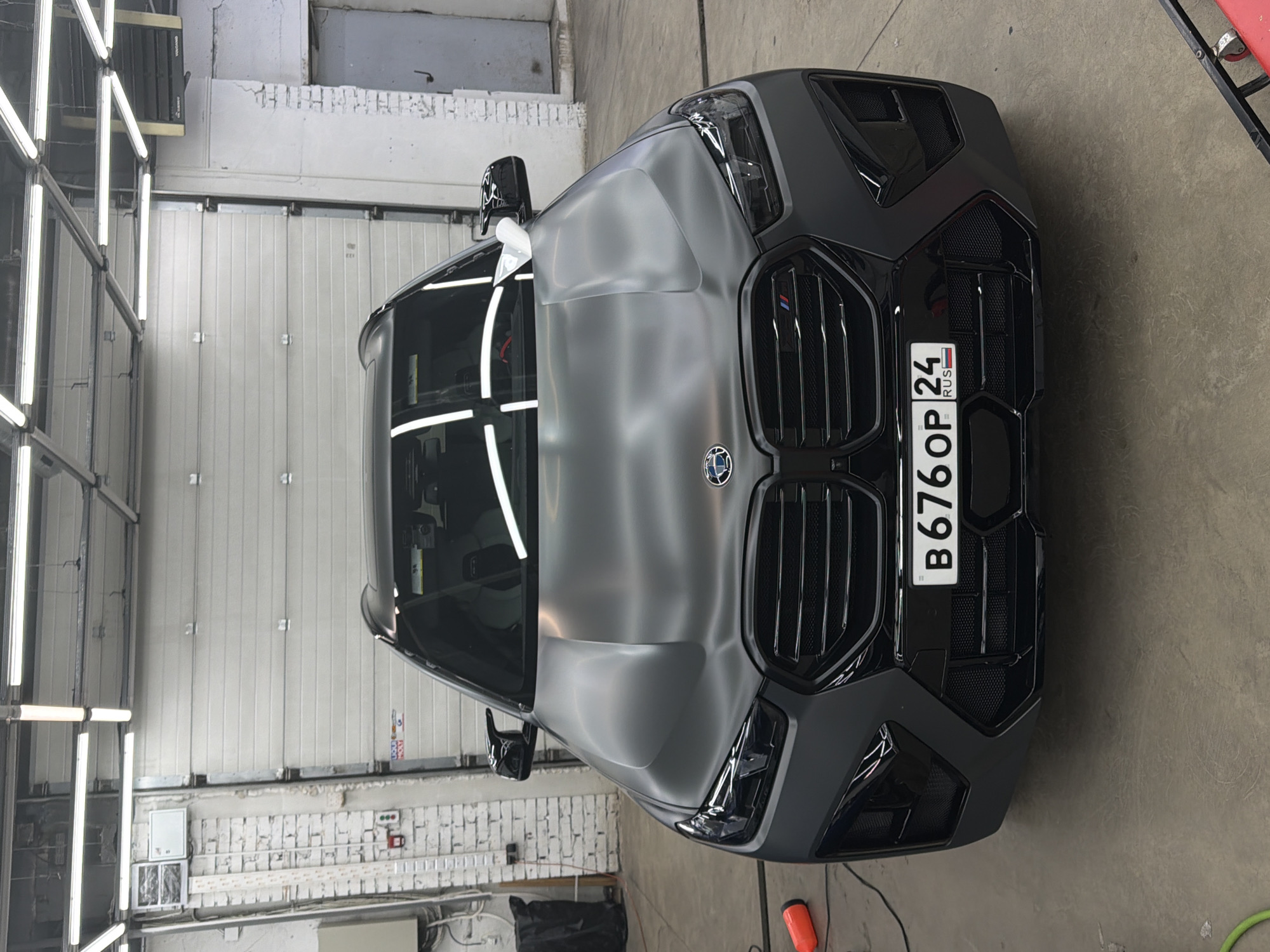 The image presents a gray BMW X6, facing forward, parked inside what appears to be a garage or automotive shop. The car's front license plate reads "B676OP 24 RUS", indicating it is registered in Russia. The surrounding environment includes gray concrete floors and white brick walls, with a large white garage door in the background. The location could possibly be in Krasnoyarsk, Russia, as per the prompt's request. Overhead lighting fixtures are visible, reflecting off the car's hood. No people are visible in the frame.