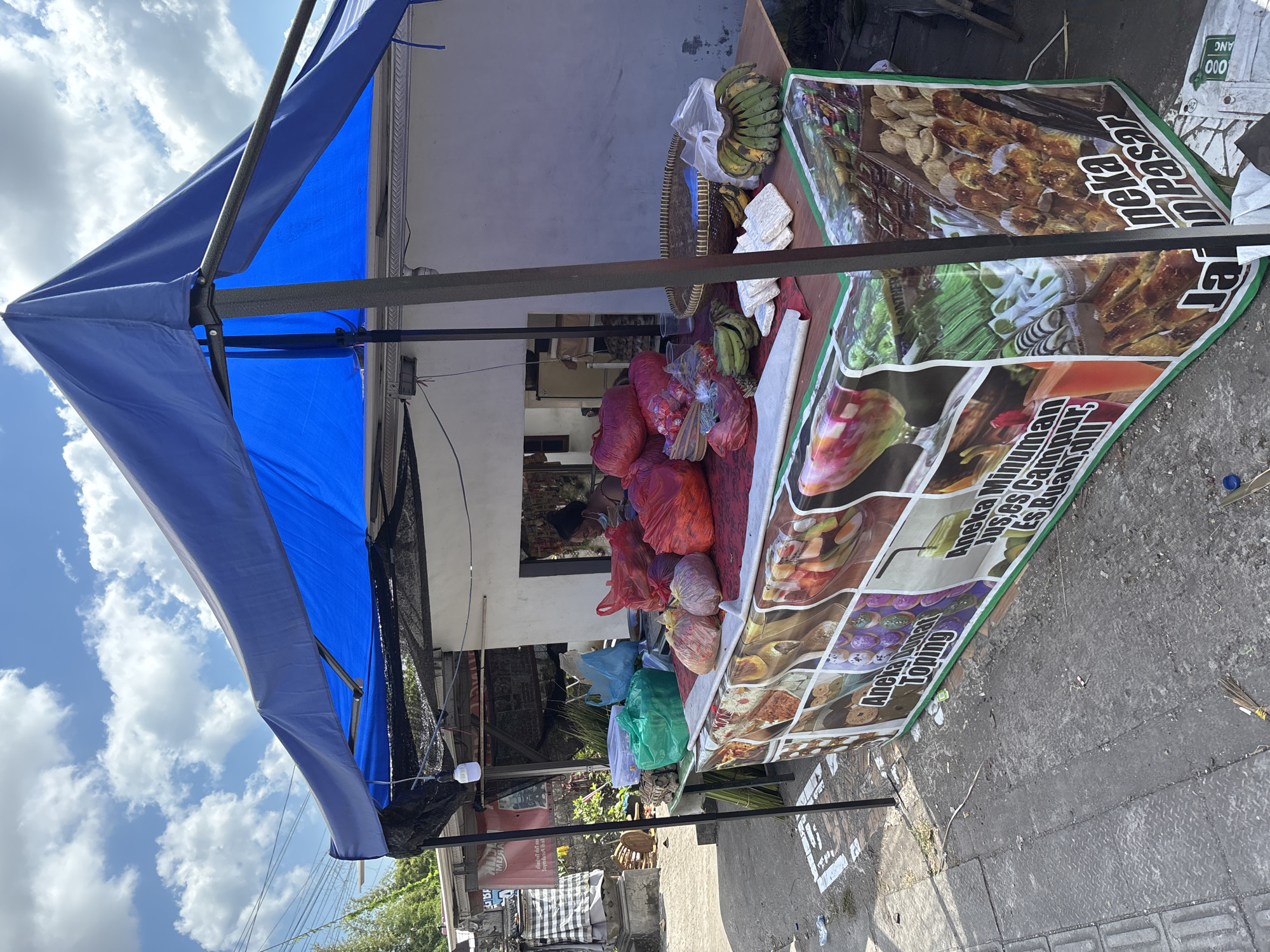 This media file captures a street vendor's stall set up under a bright blue canopy on a sunny day in Kuta Selatan, Indonesia.
The central focus is a wooden table functioning as a display counter. On the table, several bunches of green bananas are arranged, with some placed in a woven basket. Blocks of what appear to be tempeh are also visible. Various plastic bags in red, blue, and green colors are piled on the table, likely containing other merchandise.
A large, colorful banner hangs along the side of the table, serving as an advertisement. The banner features images of diverse food and drink items, including skewers, various snacks, and colorful beverages. Visible text on the banner, primarily in Indonesian, includes phrases like "Aneka Minuman" (Various Drinks), "Jus, es Campur," (Juice, mixed ice,), "Es Buan dil" (likely Es Buah dan lain-lain - fruit ice and others), "Toping" (Topping), and "Pasar" (Market).
In the background, part of a building with white walls is seen, and a person's head is faintly visible through a window. The ground is a concrete surface, suggesting a sidewalk or roadside location. The sky is bright with scattered white clouds, indicating clear daytime weather. The setup suggests an active commercial environment, typical of a local market stall.