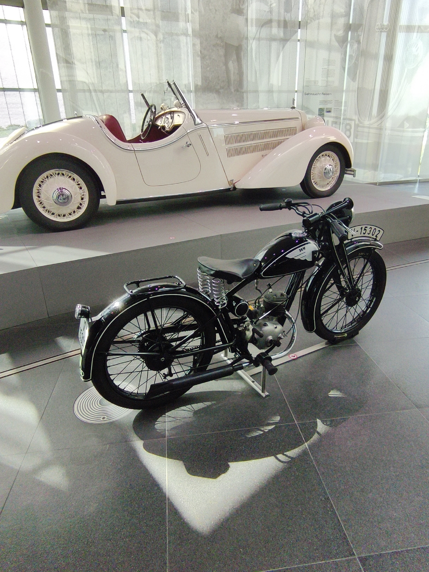 The image, taken at the BMW Museum in Ingolstadt, Germany, depicts a vintage black motorcycle in the foreground, positioned in front of a cream-colored classic convertible automobile. The motorcycle is a DKW model, evidenced by the lettering on its fuel tank. It features a single seat, spoked wheels, a visible engine, and a license plate displaying "M-15302". The car, likely from the 1930s, has a red interior and wire-spoke wheels. Both vehicles are presented on a dark, textured floor, with soft lighting casting shadows. In the background, large windows and reflective surfaces suggest an indoor exhibition space.
