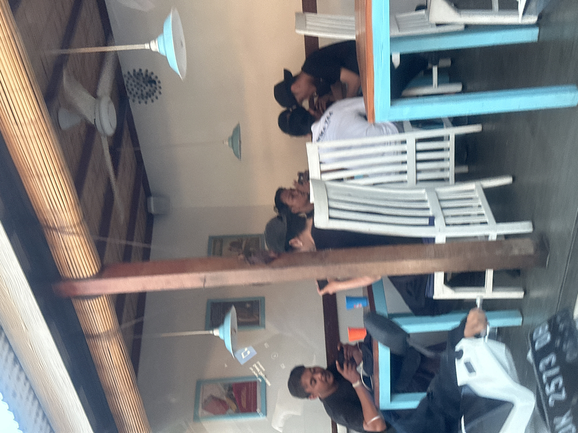 This image depicts an indoor scene, possibly a cafe or restaurant, with a rustic, open-air feel, set in Kuta, Indonesia. Several people are visible, seated at tables and interacting. In the foreground, a man in a black t-shirt is looking down at a phone. Behind him, another person is seated, also appearing to be engaged with a phone or device. Further back, a group of people are gathered around a table with blue and white furniture. One person, wearing a black cap, is leaning over the table, appearing to talk to someone. Another person in a white t-shirt is also seated at this table. The space is characterized by natural materials like wood, evident in the ceiling beams and a woven textured wall covering. White slatted chairs and tables with light blue accents contribute to a casual, beachy aesthetic. A ceiling fan is visible in the upper left portion of the image, suggesting a warm climate. The lighting appears to be natural, possibly from overhead or from unseen windows, indicating it's daytime. The overall mood seems relaxed and social. Visible text includes a license plate on the right side, which reads "DK 2573 DB".