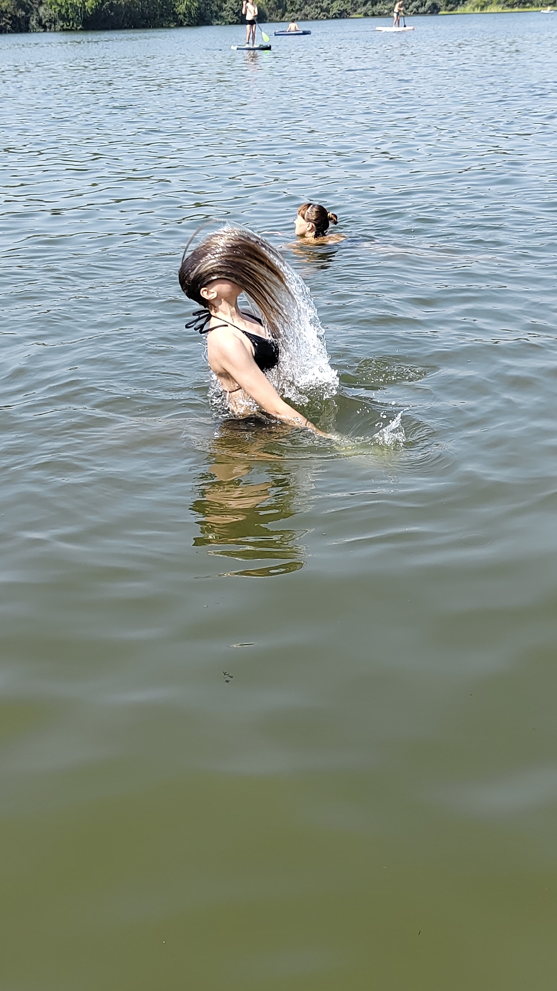 In Terent'yevo, Russia, on a body of water, a woman in a black bikini is in the water, with her head bent backward and her long, wet hair splashing outwards, creating a wave of water. Another person is visible in the water behind her. In the distance, on paddleboards in the water, are two individuals. The environment is a sunny day with trees visible on the far bank of the water.
