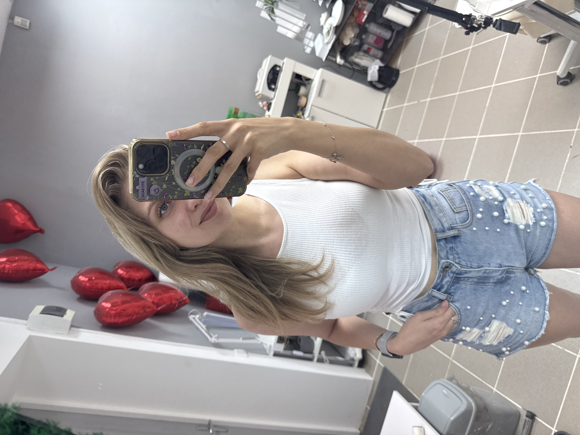 A woman is taking a selfie indoors. She is wearing a white ribbed tank top and light blue denim shorts with faux pearl embellishments and distressed details. She is holding a smartphone in her right hand, obscuring part of her face. The phone has a decorative case with floral patterns. She is also wearing a ring on her finger and a watch.
In the background, several red heart-shaped balloons are visible. A white shelving unit or table is also in the frame, along with other miscellaneous objects. The location appears to be indoors, with a tiled floor.
There is no information available to confirm the location as Izhevsk, Russia.