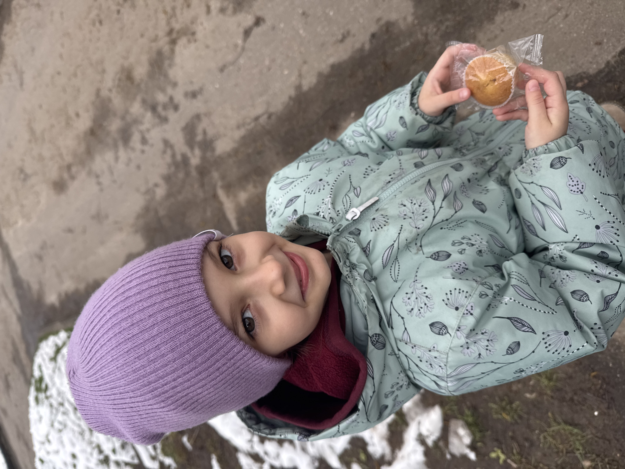 A high-angle, tilted photograph captures a young child, likely a girl, outdoors in Раздолье, Russia. The child is wearing a bright purple knit beanie and a light mint-green winter jacket featuring a delicate pattern of white and black leaves and abstract floral designs. A maroon fleece lining is visible around her neck. She looks up towards the camera with a bright, joyful smile. Her hands are gently holding a single, round muffin or cupcake, still enclosed in its clear plastic packaging and paper liner. The ground beneath her is a textured mix of brown earth or concrete, interspersed with patches of melting snow, indicating cold weather, likely late winter or early spring. The lighting is soft and natural, suggesting an overcast daytime. There is no visible text in the image.