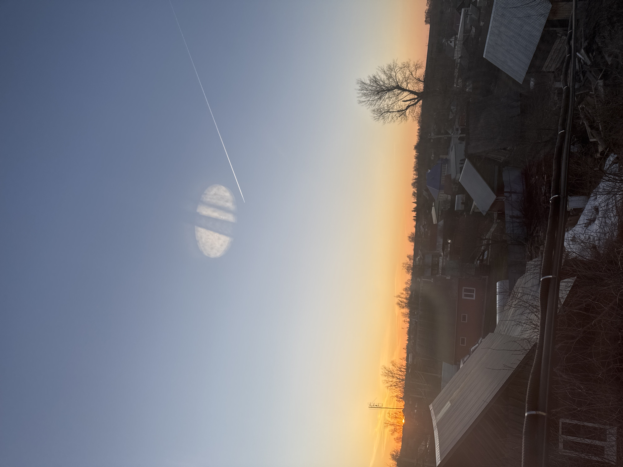 The image shows a landscape in Фоки, Russia. The sky transitions from blue to orange, indicating either sunrise or sunset. An airplane contrail cuts diagonally across the sky. There is a circular lens flare near the contrail. The horizon features silhouettes of trees, buildings, and rooftops. Power lines are visible in the foreground. A leafless tree stands out against the orange sky.