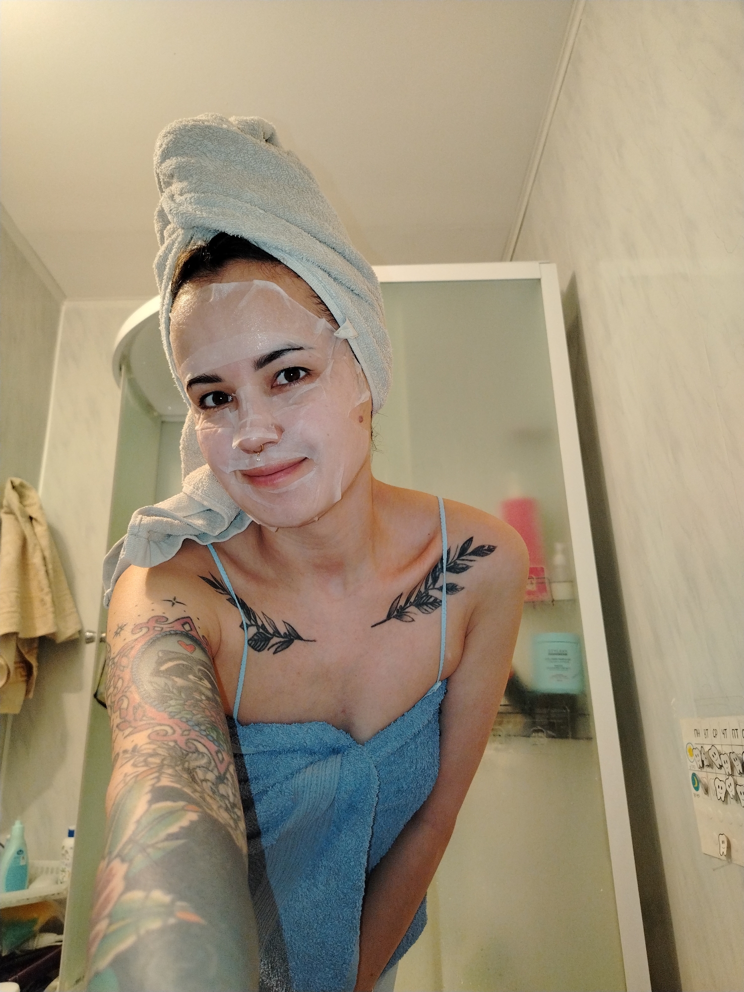 A person is in a bathroom, wearing a white sheet mask on their face and a light blue towel wrapped around their body. A light grey towel is wrapped around their head. The person has tattoos on their left arm and chest. The bathroom has light-colored walls, a shower, and some toiletries. A tan towel hangs in the background. A child's magnetic calendar is stuck to the wall of the shower. The location is likely in a residential bathroom, possibly in an apartment or house. The city of Noviy, Russia is not evident in the image.