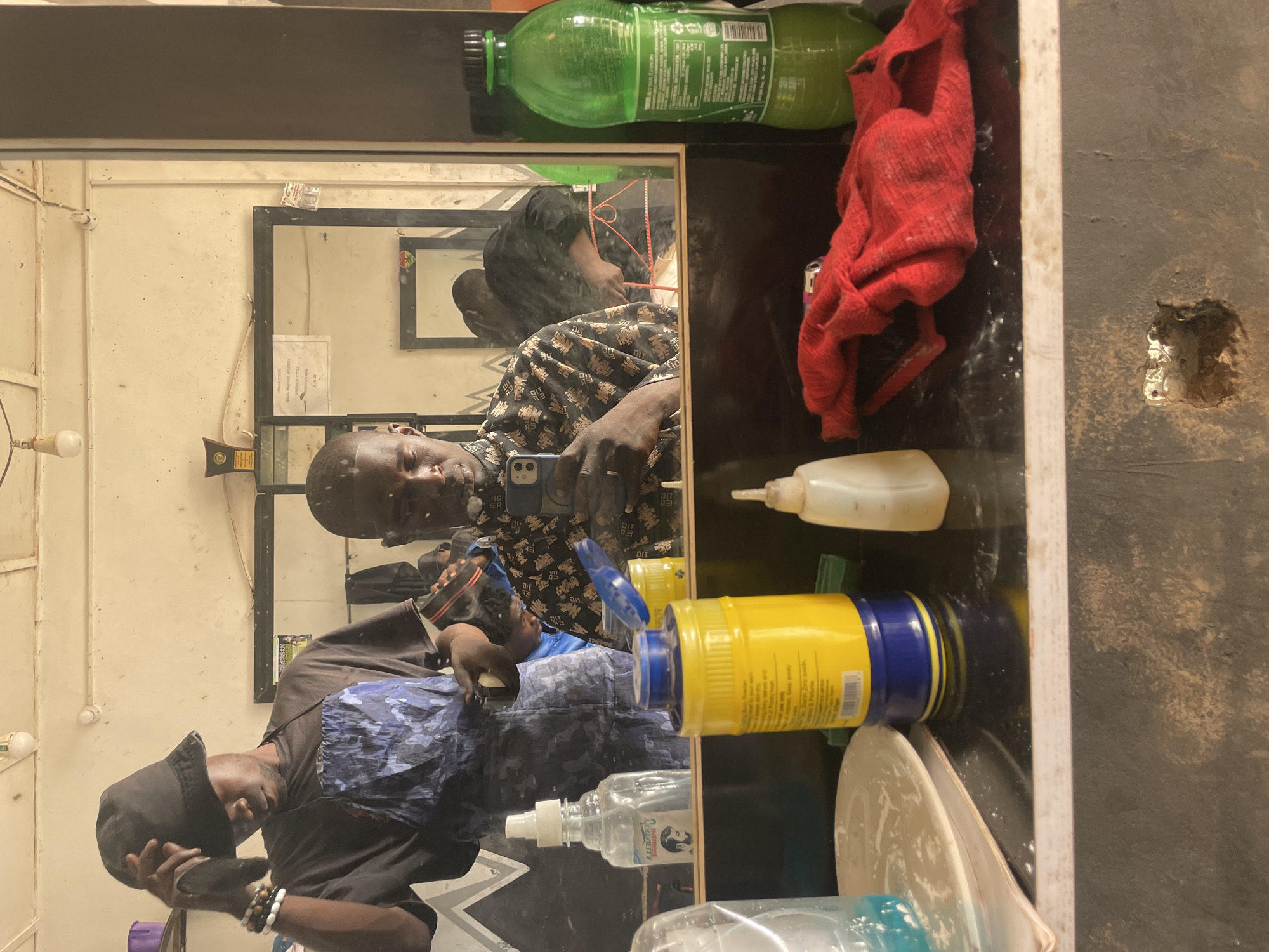 A person is seen in the mirror with another person cutting their hair. The image is taken in what appears to be a barbershop, with various barbering supplies present. A water bottle and other objects are on the counter next to the mirror. The condition of the walls and surroundings suggests a modest setting. While the image is taken in Durimi, Nigeria, there is no additional information available about the precise location.
