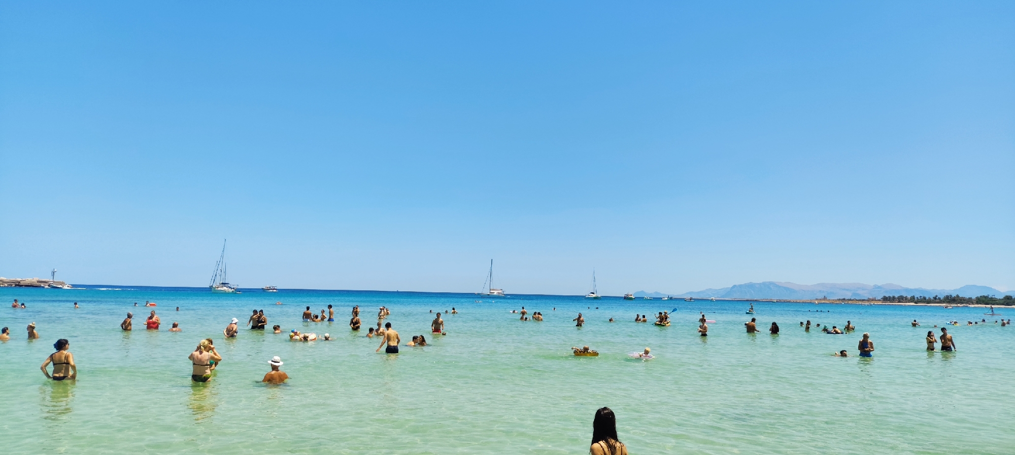 This is a wide, panoramic, eye-level, outdoor, daytime, full-shot photograph of a crowded beach scene at San Vito Lo Capo, Italy. The water is clear and turquoise, with a sandy bottom visible in shallower areas. The sky is a bright, cloudless blue. In the foreground, a woman with dark hair is facing away from the camera, standing in the water. Scattered throughout the water are dozens of people swimming, wading, and relaxing. Some are in groups, while others are alone. There are several inflatable toys and floats in the water. In the middle ground, numerous sailboats and smaller motorboats are anchored or cruising. The coastline in the background features a mountainous landscape with sparse vegetation and buildings. The overall impression is one of a lively and popular beach on a sunny day.