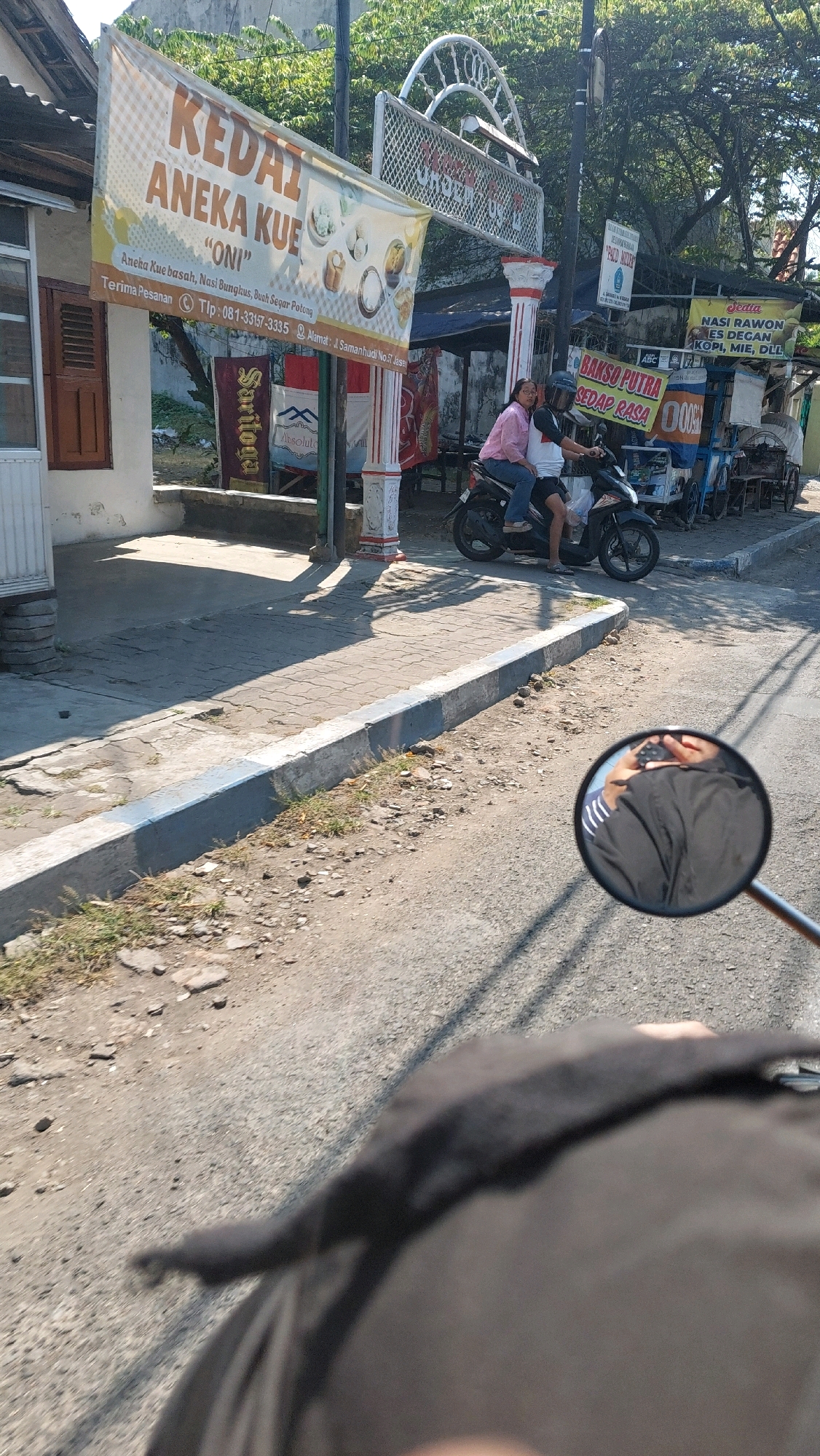 In Sidoarjo, Indonesia, on a sunny day, a motorcycle is parked in front of a shop. Two people, a woman in a pink shirt and a man in a white shirt, are seated on the motorcycle, appearing to be resting or waiting. The woman is looking towards the side, while the man is looking ahead. Behind them, a banner for "KEDAI ANEKA KUE 'ONI'" is displayed, advertising various cakes and snacks. The shop itself is adorned with a decorative white archway. A street runs in front of the shop, with a paved sidewalk. In the foreground, a motorcycle's rearview mirror reflects the scene, showing the rider's arm and shoulder. The environment is characterized by a mix of urban and natural elements, with buildings, trees, and a clear blue sky. The overall atmosphere suggests a typical day in a small town or city area.