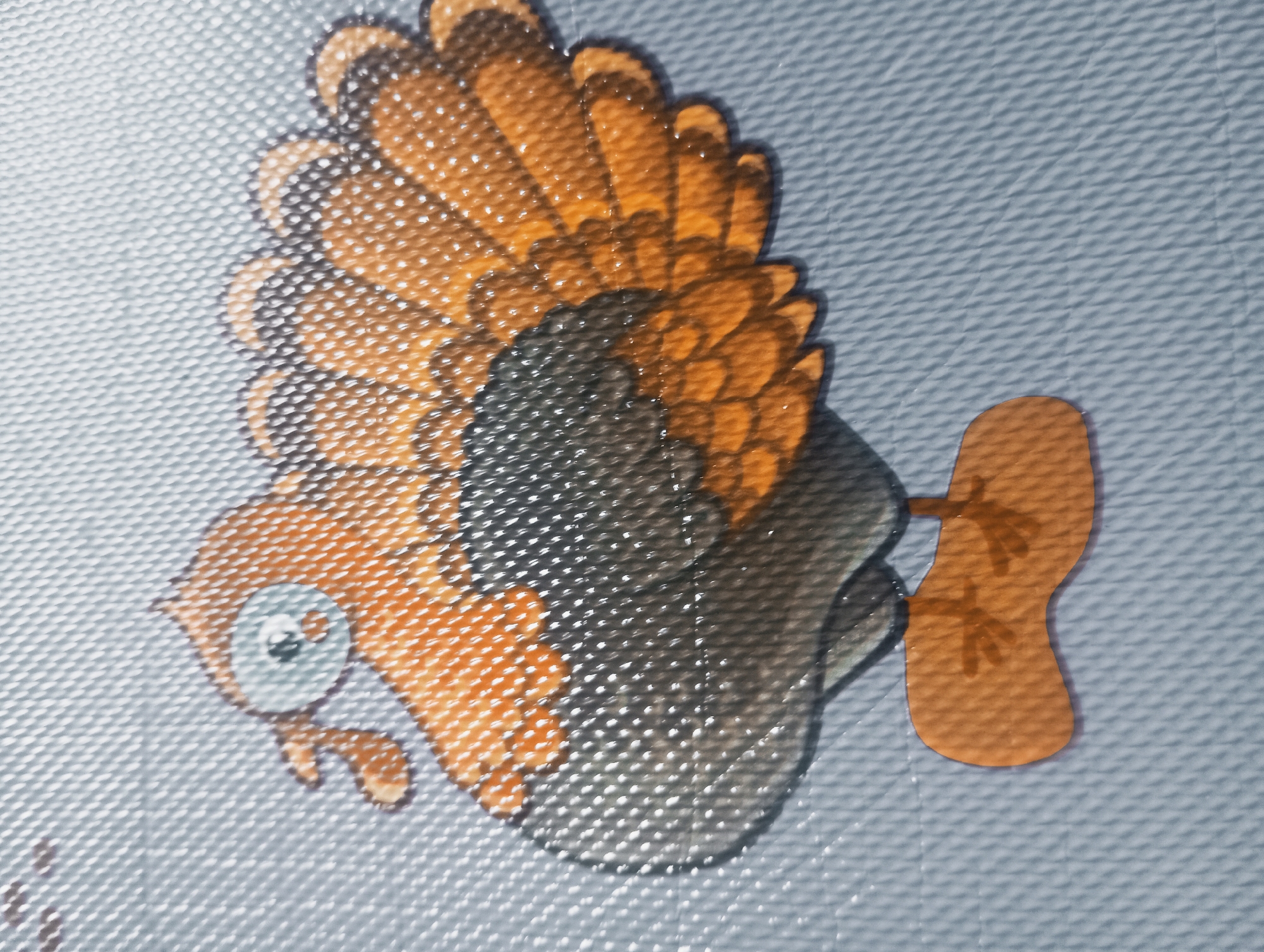 The image is a close-up, eye-level, medium shot of a cartoonish turkey depicted on a light blue, textured surface. The turkey is predominantly orange and brown with grey accents on its body and tail feathers. It has a large, circular, light blue eye with a black pupil and a small orange beak. The turkey is standing on a flat, orange base with two visible toes. The texture of the surface suggests it might be a mat or a floor covering. The image is well-lit, and there are no people or other discernible objects or environmental features in the frame. The specific location within Kutasari, Indonesia, cannot be determined from the visual information provided.