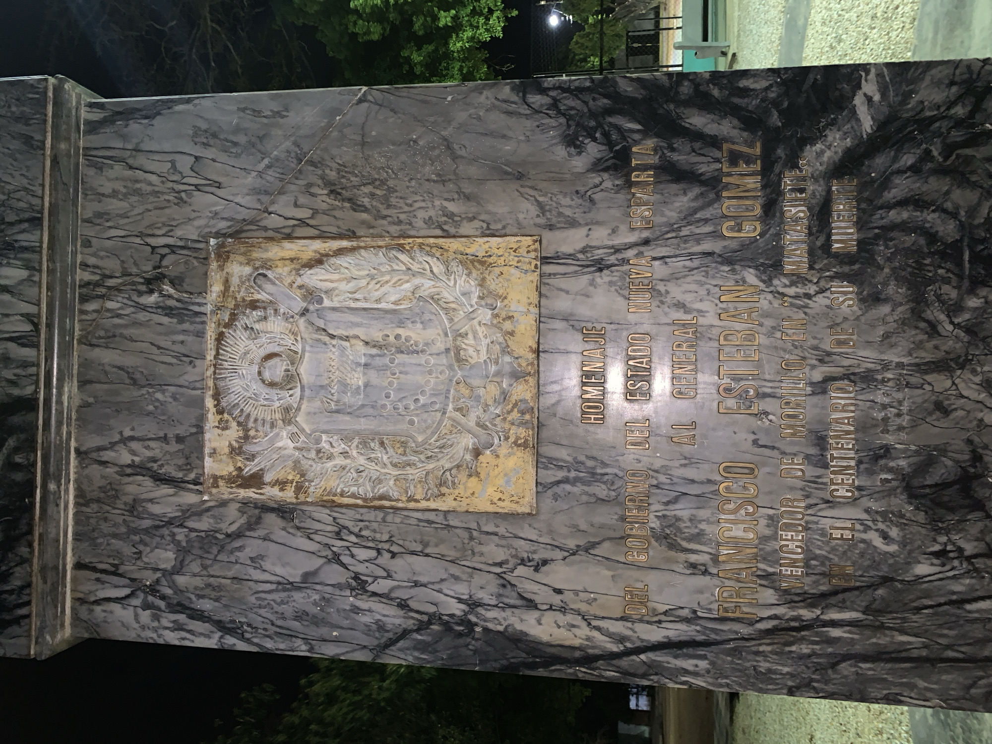 The image displays a commemorative monument, likely a plinth or large plaque, made of dark, veined marble. Attached to the marble is a rectangular, aged metal plaque featuring an ornate, relief-sculpted emblem, possibly a coat of arms, showing signs of tarnish and wear.
Inscribed on the marble, in gold-colored capital letters, is text in Spanish. The inscription reads:
"HOMENAJE
DEL GOBIERNO DEL ESTADO NUEVA ESPARTA
AL GENERAL
FRANCISCO ESTEBAN GOMEZ
VENCEDOR DE MORILLO EN MATASIETE
EN EL CENTENARIO DE SU MUERTE"
This translates to:
"HOMAGE
FROM THE GOVERNMENT OF NUEVA ESPARTA STATE
TO GENERAL
FRANCISCO ESTEBAN GOMEZ
VICTOR OF MORILLO AT MATASIETE
ON THE CENTENNIAL OF HIS DEATH"
The monument honors General Francisco Esteban Gómez, celebrating his victory over Morillo at Matasiete. The commemoration took place on the centennial of his death.
The scene appears to be at night, with artificial lighting illuminating the monument. The background is dark, with silhouettes of trees visible, and hints of a paved area or building. The monument is situated in Santa Ana, Venezuela, indicating its historical and regional significance. The overall impression is one of solemn remembrance and historical tribute.