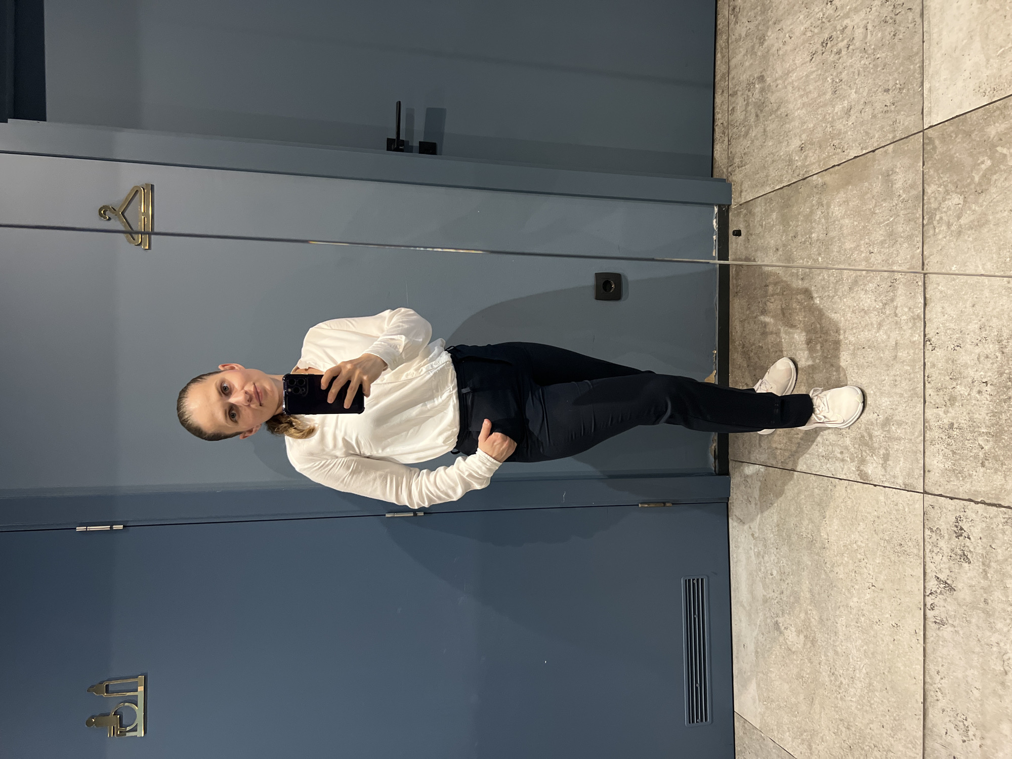 A woman stands in a changing room, taking a selfie in a mirror. She is wearing a white long-sleeved shirt and dark trousers, with white sneakers. The changing room has blue walls and a tiled floor. There are signs for a disabled toilet and a clothes hanger on the wall. A power outlet is visible near the floor. The overall impression is of a practical and functional space. This location is likely a fitting room within a retail store in Budva, Montenegro.