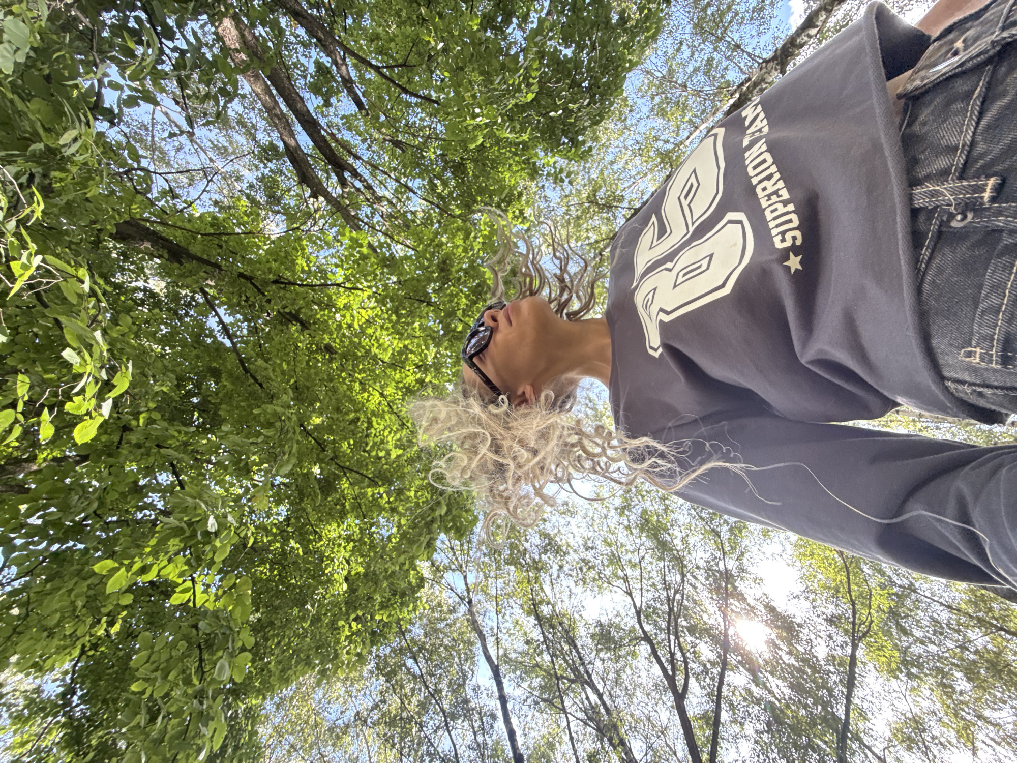 An outdoor, low-angle, eye-level shot captures a person from the chest up, looking upwards towards a canopy of lush green trees and the bright blue sky. The person, positioned in the lower right of the frame, has blonde, curly hair that is windswept. They are wearing large, black, rectangular sunglasses and a dark grey long-sleeved shirt with a white graphic on the front that appears to say "85" and "SUPERIOR TEAM." The person is also wearing dark denim jeans. The sun shines through the leaves, creating a dappled light effect across the scene. The branches of the trees create a natural frame at the top and sides of the image. There are no recognizable landmarks indicating the specific location in Rusanovo, Russia.