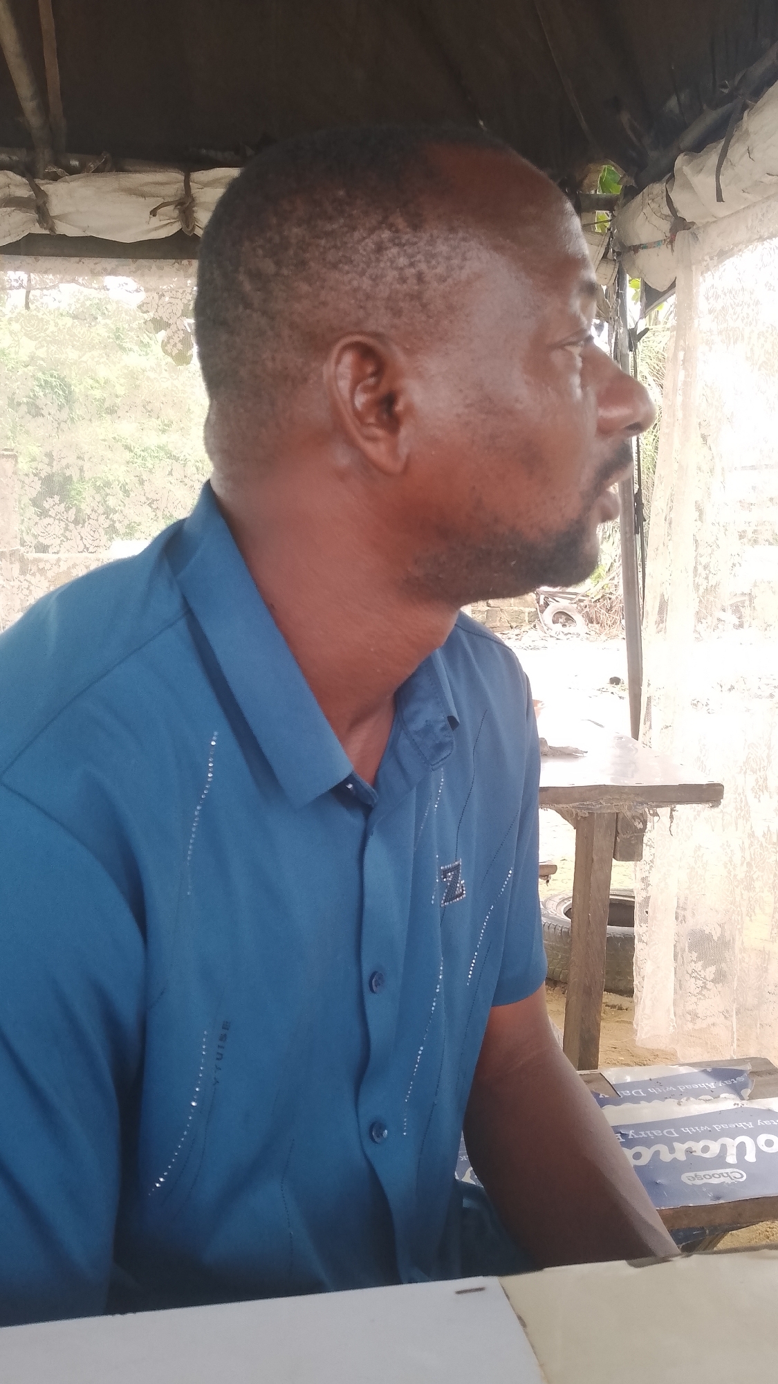 The image shows a middle-aged Black man with short, dark hair and a goatee, sitting at a table outdoors. He is wearing a blue, short-sleeved button-down shirt with decorative elements. He is looking to his right. The setting appears to be a covered outdoor space, possibly a roadside eatery or shelter. There is a table visible, and behind it, objects that suggest an automotive repair shop or storage area with tires and other equipment. The location is indicated as Effurun, Nigeria, a city near Warri. The background includes a translucent curtain-like barrier. The man is positioned in front of a table covered with a promotional sign for dairy products.