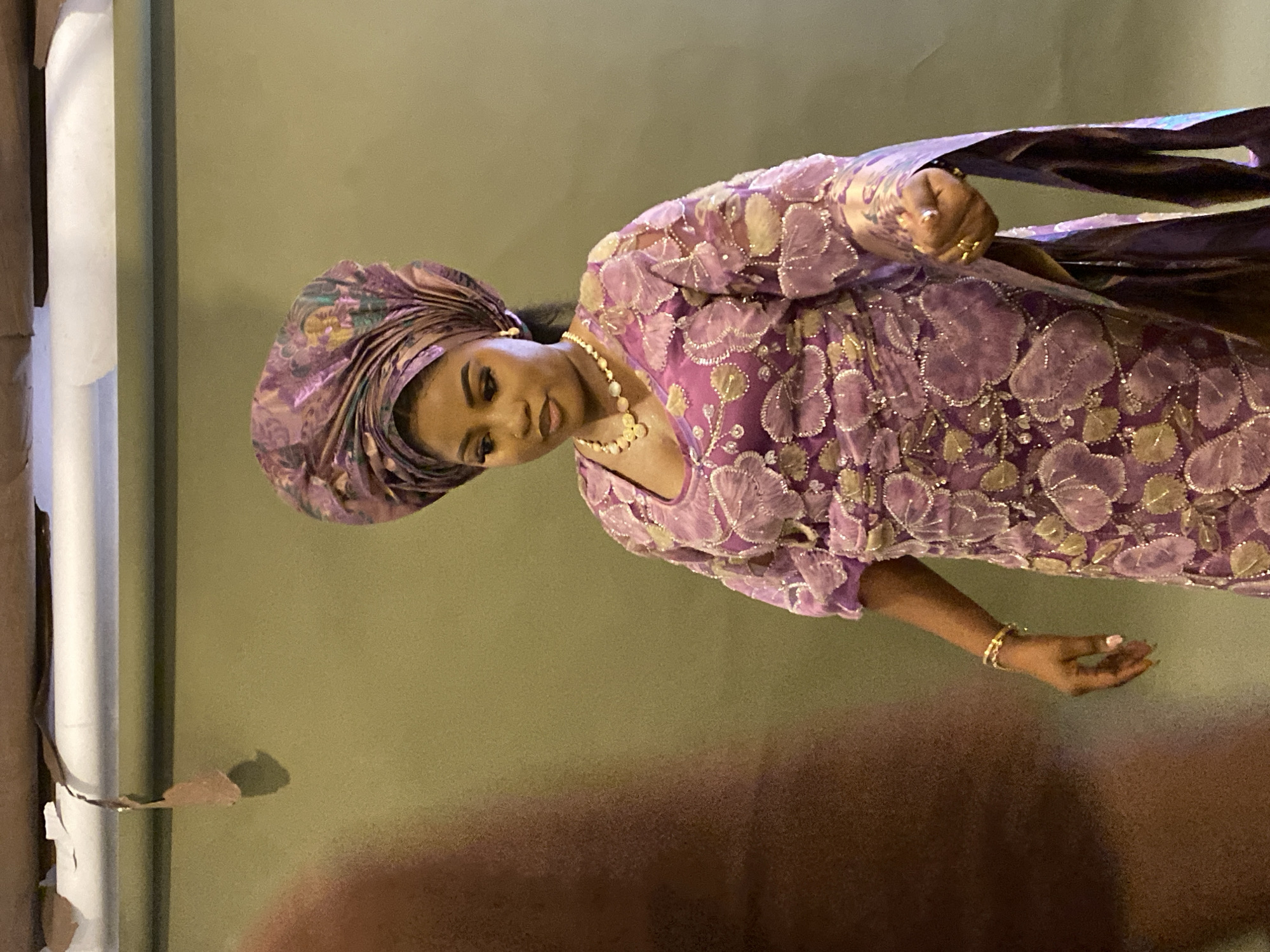 The image displays a woman posing indoors, elegantly dressed in traditional West African attire, consistent with Nigerian fashion from Badagry, Nigeria. She wears a vibrant purple ensemble, featuring an intricately tied headwrap (gele) and a matching dress adorned with elaborate floral patterns in gold and silver embroidery or lace. Her accessories include a multi-strand pearl necklace, earrings, and two gold-toned bracelets on her left wrist. Her fingernails are manicured with a light pink polish.
The woman is positioned slightly to the left of the frame, facing the camera with a serene expression and a gentle smile, her head tilted subtly. Her right hand is raised, delicately holding a piece of flowing purple fabric, while her left hand rests by her side.
The setting appears to be indoors, against a plain, light olive-green wall. To the far left, a vertical white structure, possibly a pole or column, is visible adjacent to a brown, textured surface. The lighting is soft and warm, suggesting artificial indoor illumination. There is no visible text within the image.