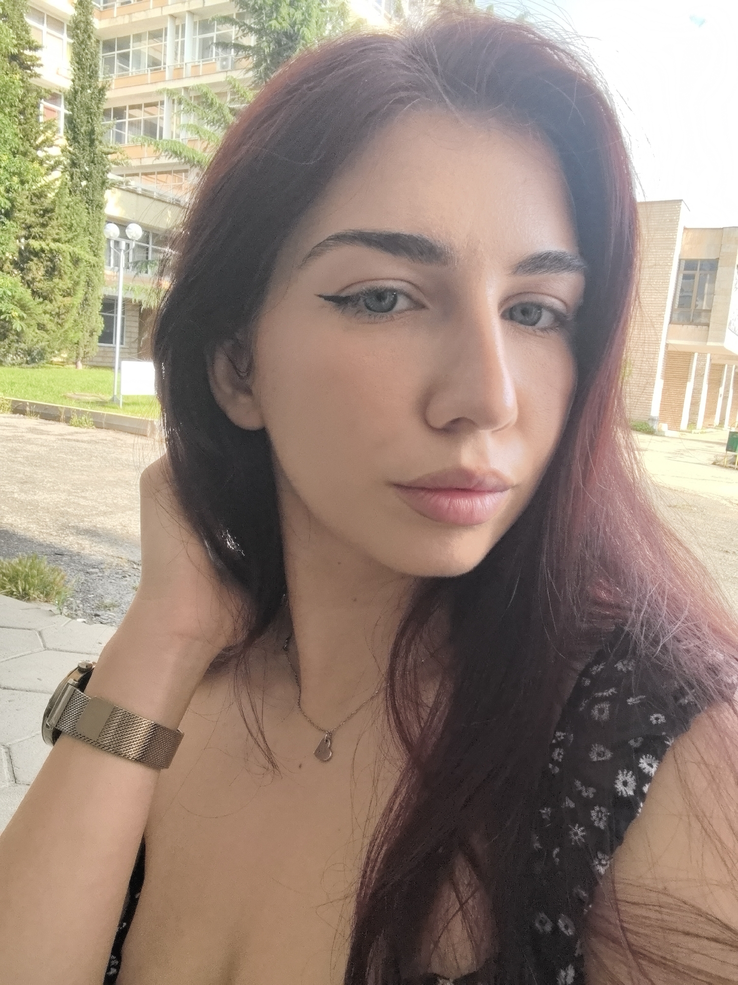 A young woman is captured in a head-and-shoulders selfie, positioned outdoors in Bogomilovo, Bulgaria. She has long, dark reddish-brown hair, styled loosely, and light blue-grey eyes accentuated by winged eyeliner. Her eyebrows are well-defined, and her lips have a natural, soft pink color. She wears a dark top, possibly a dress, featuring a subtle white floral or patterned print. Her accessories include a delicate silver-toned necklace with a small heart-shaped pendant and a metallic, possibly gold or bronze-toned, mesh-strap watch on her left wrist. She holds her left hand near her head, a common selfie pose, and gazes directly at the camera with a neutral expression.
The background depicts a bright, sunny daytime environment. Behind her stand several multi-story buildings, characterized by numerous windows and balconies, suggesting an institutional or older residential architectural style. Tall, slender evergreen trees and other green foliage, including bushes and grass, are visible. A lamppost can be seen in the middle ground. The ground is a mix of paved areas and natural terrain, with strong shadows indicating clear weather. The setting appears to be an urban or campus-like area. No visible text is present in the image.