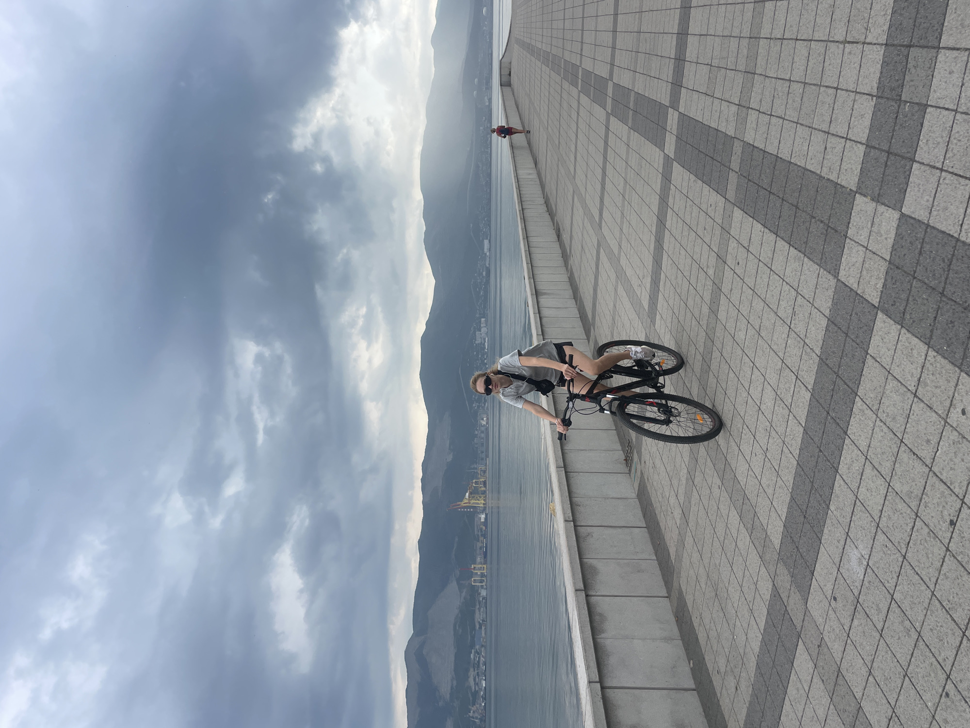 In Novorossiysk, Russia, a woman is captured mid-cycle on a paved promenade adjacent to a body of water. The promenade is laid with a repeating pattern of square tiles in light and dark gray. A concrete barrier separates the promenade from the water, which stretches towards a mountainous horizon. The sky above is overcast with heavy, gray clouds, intermittently illuminated by rays of light piercing through. In the background, distant industrial structures with cranes are visible on the shoreline. Further along the promenade, a person in red is seen running. The woman on the bicycle wears a gray t-shirt, black shorts, sunglasses, and a dark crossbody bag. She is actively engaged in cycling, with her gaze directed forward.