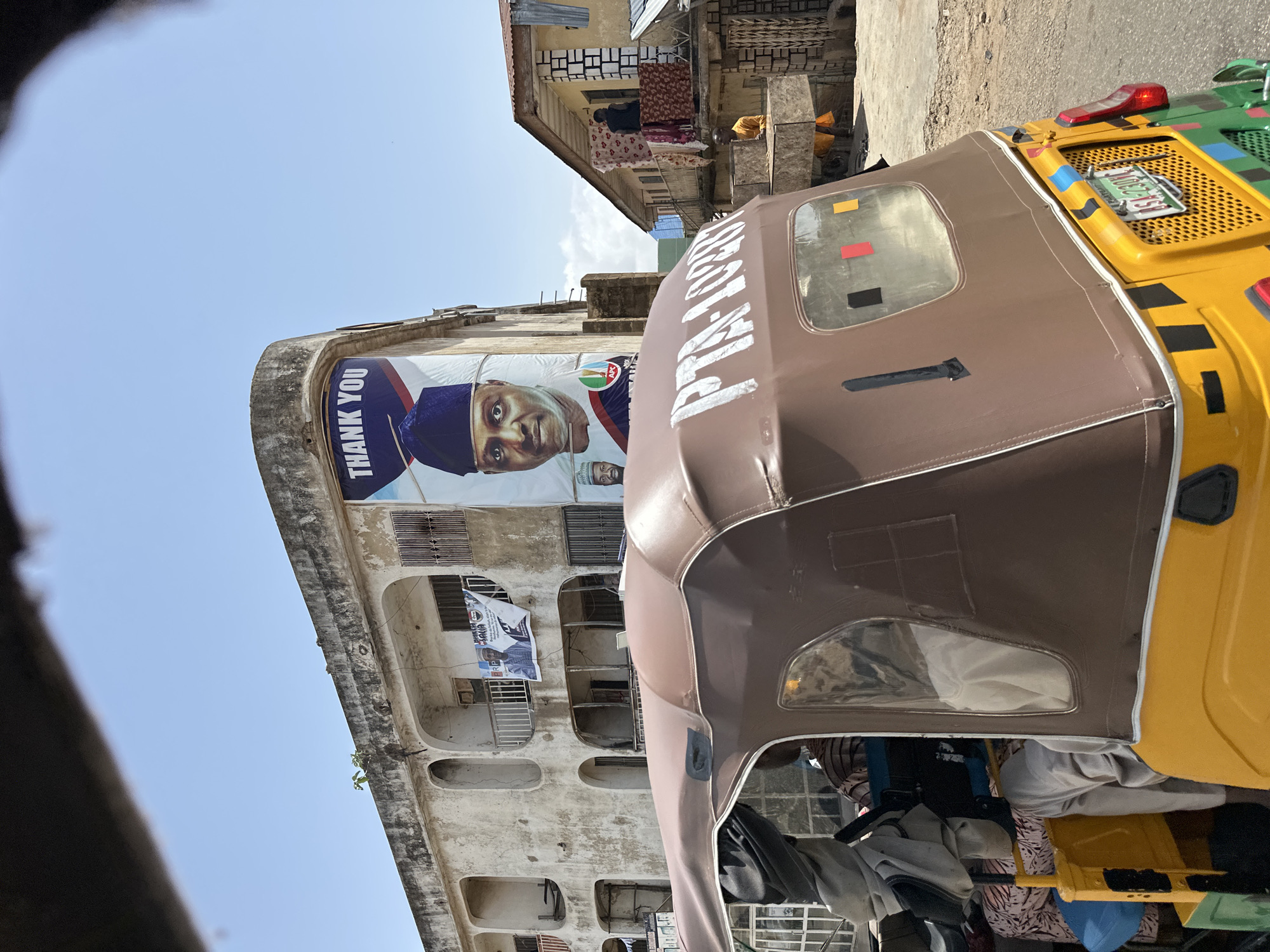 In Jos, Nigeria, a yellow and green auto-rickshaw with a brown canvas top is parked on a paved street. The auto-rickshaw has the number "PTA-1020" written in white on its roof and a license plate that reads "ASA 2309 A". In the background, a weathered, light-colored building with multiple balconies and arched openings stands. A large banner featuring a portrait of a man in a blue cap and the text "THANK YOU" is displayed on the side of the building. Another smaller banner with two men is visible on one of the balconies. Adjacent to this building is another structure with a corrugated metal roof and balconies where people are present, including one individual standing on a balcony with laundry hanging. The environment appears to be an urban setting with a clear sky.