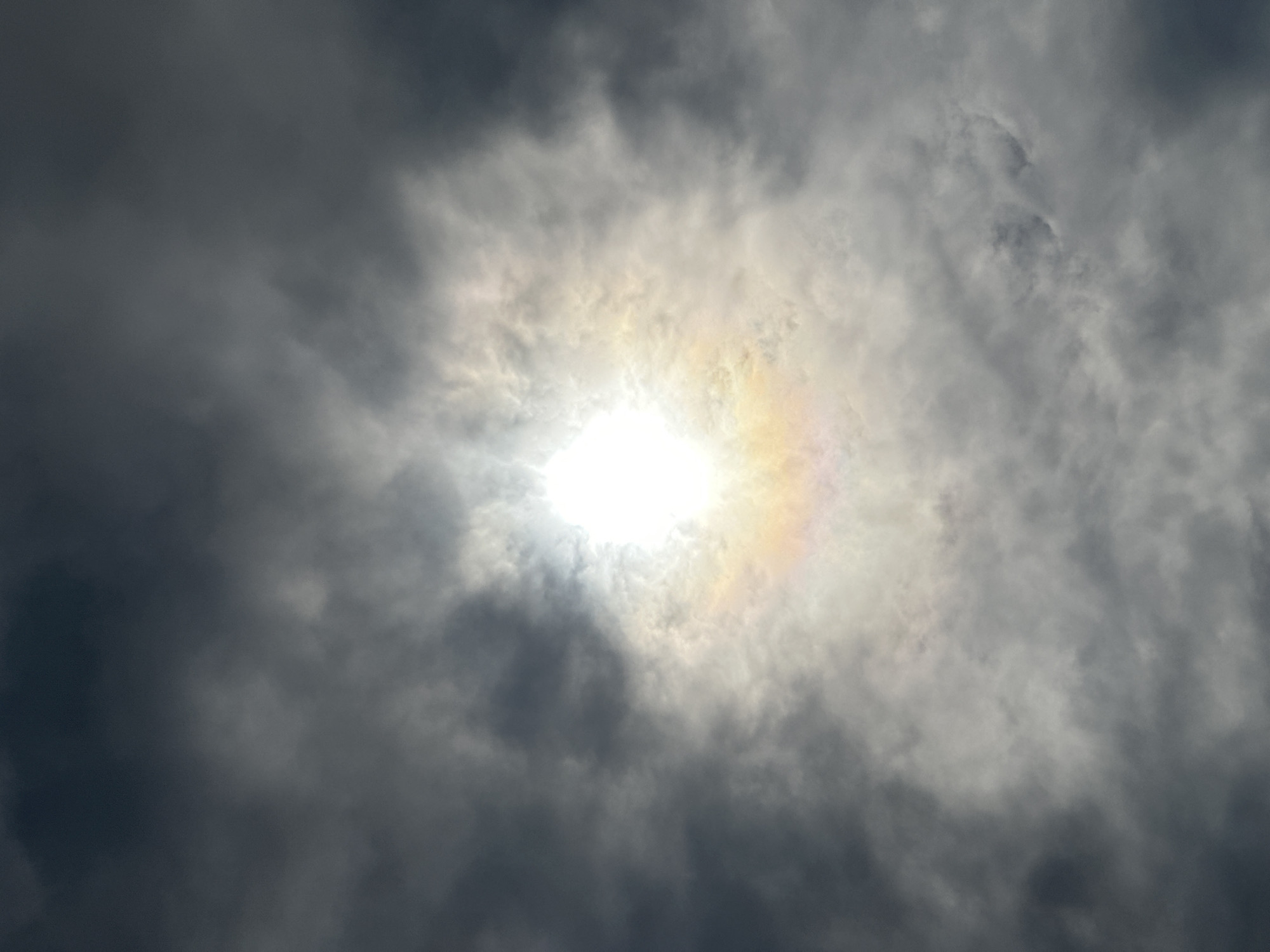 The image shows a bright sun shining through a cloudy sky. There is a visible halo around the sun, with a faint rainbow of colors. The clouds are a mix of light and dark gray shades. The image was taken in Nizhny Novgorod, Russia.