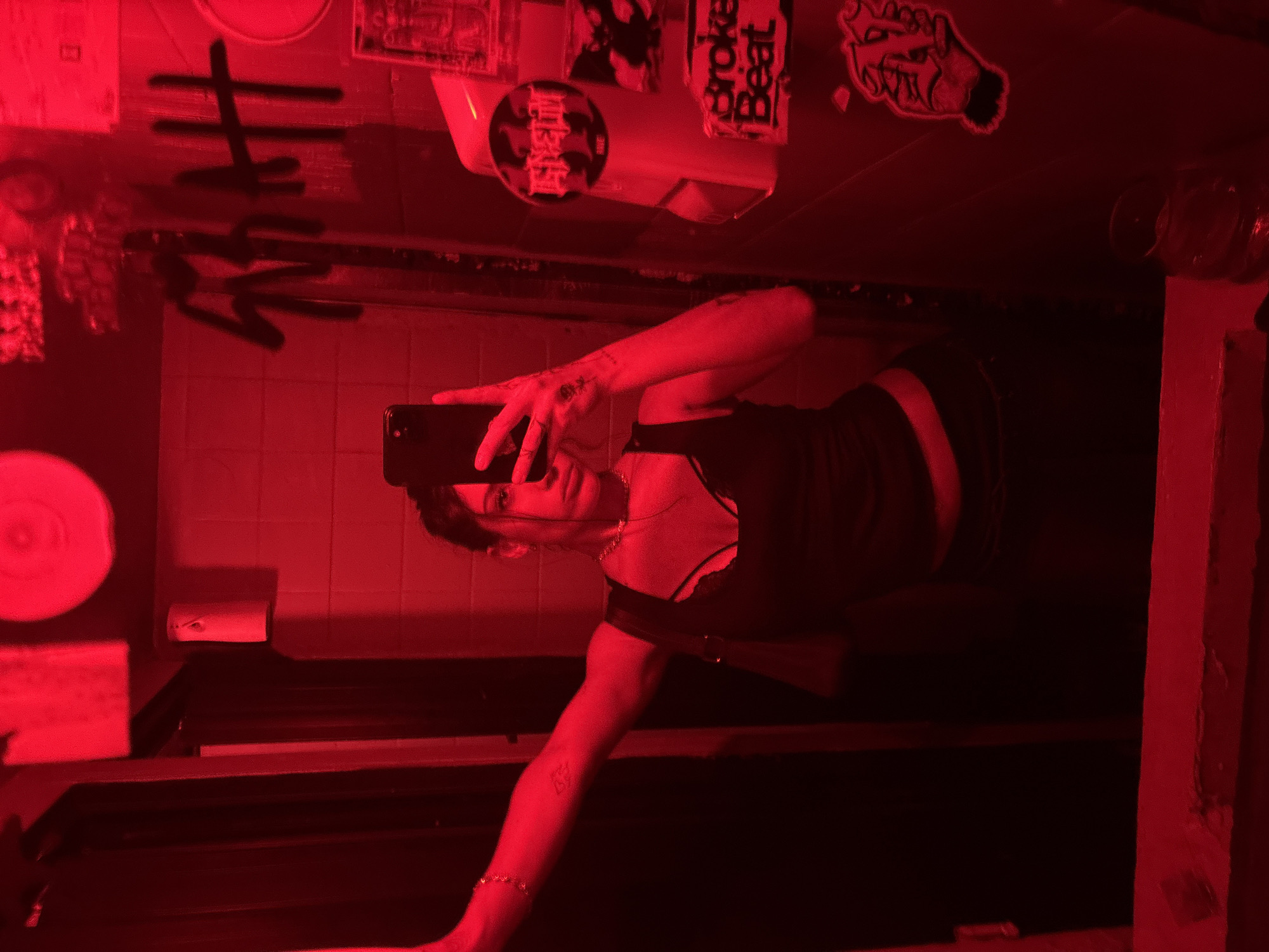 A person takes a mirror selfie in a red-lit bathroom. The subject is holding a smartphone and is wearing a dark top. Behind them, the wall has tiles, and there are various stickers and graffiti. A toilet paper dispenser and toilet can be seen. The location appears to be a bathroom in a public space, potentially a nightclub or bar. The location in Buenos Aires, Argentina is unknown.