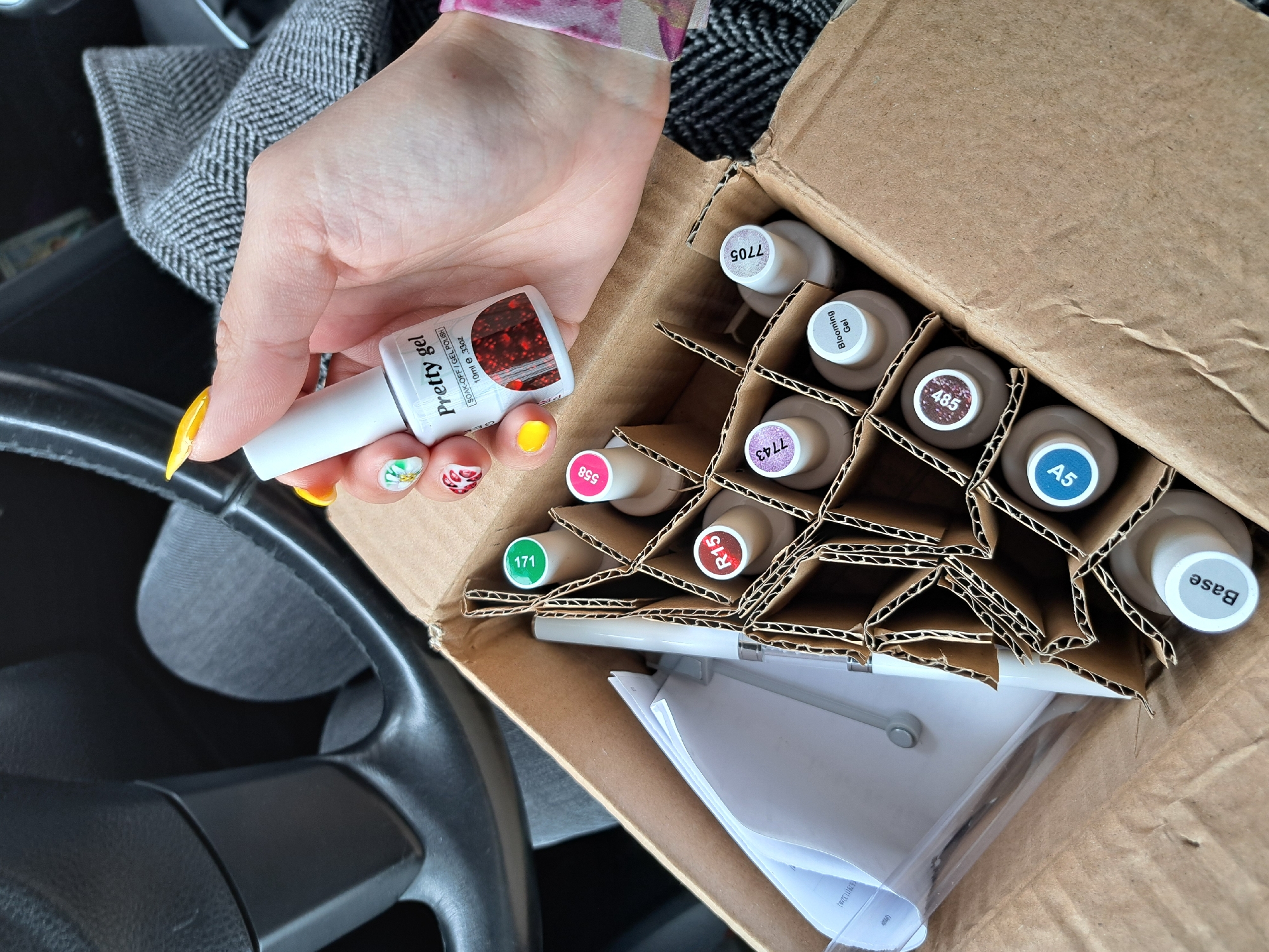 A close-up shot shows a person's hand holding a bottle of "Pretty gel" nail polish above a box filled with similar nail polish bottles. The bottles are organized in a cardboard divider. Labels on the tops of the bottles show numbers and names, including "Base", "A5", "485", "R15", "7705", "171", "558", "7743" and "Blooming Gel". The hand holding the nail polish has painted fingernails with different colors and designs. Part of a black steering wheel can be seen at the bottom left of the frame, suggesting the scene is inside a car. The gray interior of the car and what appears to be a jacket can also be seen. The location is indiscernible without additional contextual details.