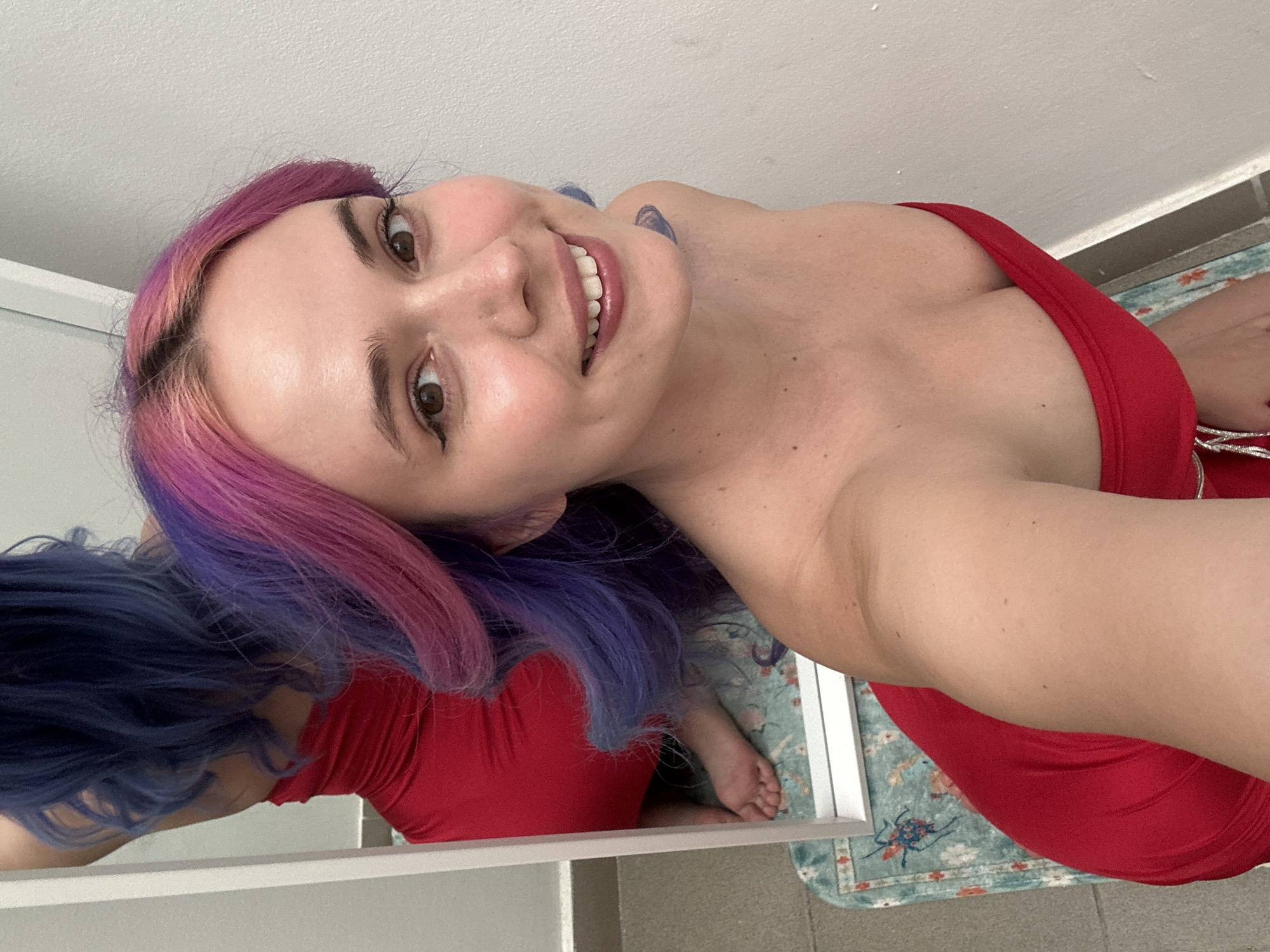 A young woman is captured in a close-up, high-angle selfie, smiling directly at the camera. Her head is tilted, and her vibrant multi-colored hair, transitioning from pink at the roots to purple and blue at the ends, frames her face. She has brown eyes, defined eyebrows, and subtle eyeliner. Her lips are slightly parted in a broad smile, revealing white teeth. A small, dark blue tattoo is visible on her right shoulder/neck area.
She is wearing a bright red strapless top or dress, with a glimpse of silver or metallic detailing visible on the right side of the garment. The background consists of a plain white wall and ceiling, suggesting an indoor setting. A mirror or reflective surface in the lower left shows her reflection from the back, revealing more of her blue hair and the red attire, as she leans forward. Below the mirror, a light blue/green patterned rug with red/orange designs (possibly insects) is visible on a tiled floor. The overall lighting appears bright, indicative of daytime. The woman's expression conveys happiness or playfulness. There is no visible text in the image. No specific visual cues confirm the location as Zagreb, Croatia, beyond the provided context.