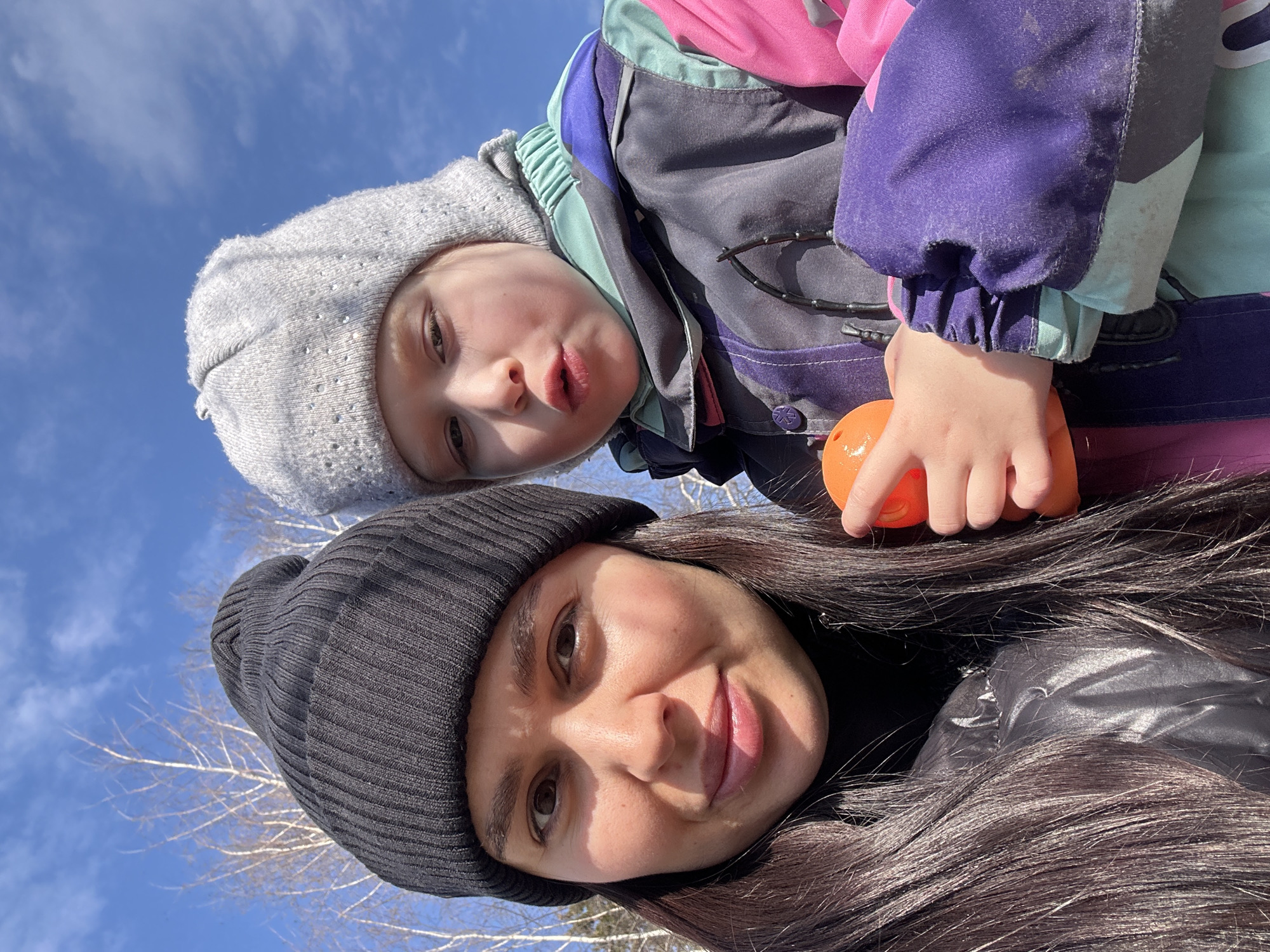 A selfie taken outdoors, likely in the city of Chaikovsky, Russia, features two individuals: an adult female and a child. The adult is in the foreground, wearing a black beanie and a black jacket. She has long, dark brown hair and is smiling. Behind her and slightly above is a young child with fair skin, wearing a grey beanie. The child is making a funny face. The child is also wearing a multicolored winter jacket. The child holds an orange toy. In the background, a bright blue sky with scattered clouds and bare tree branches are visible. The image has a slightly tilted angle, suggesting it was taken with a handheld device.