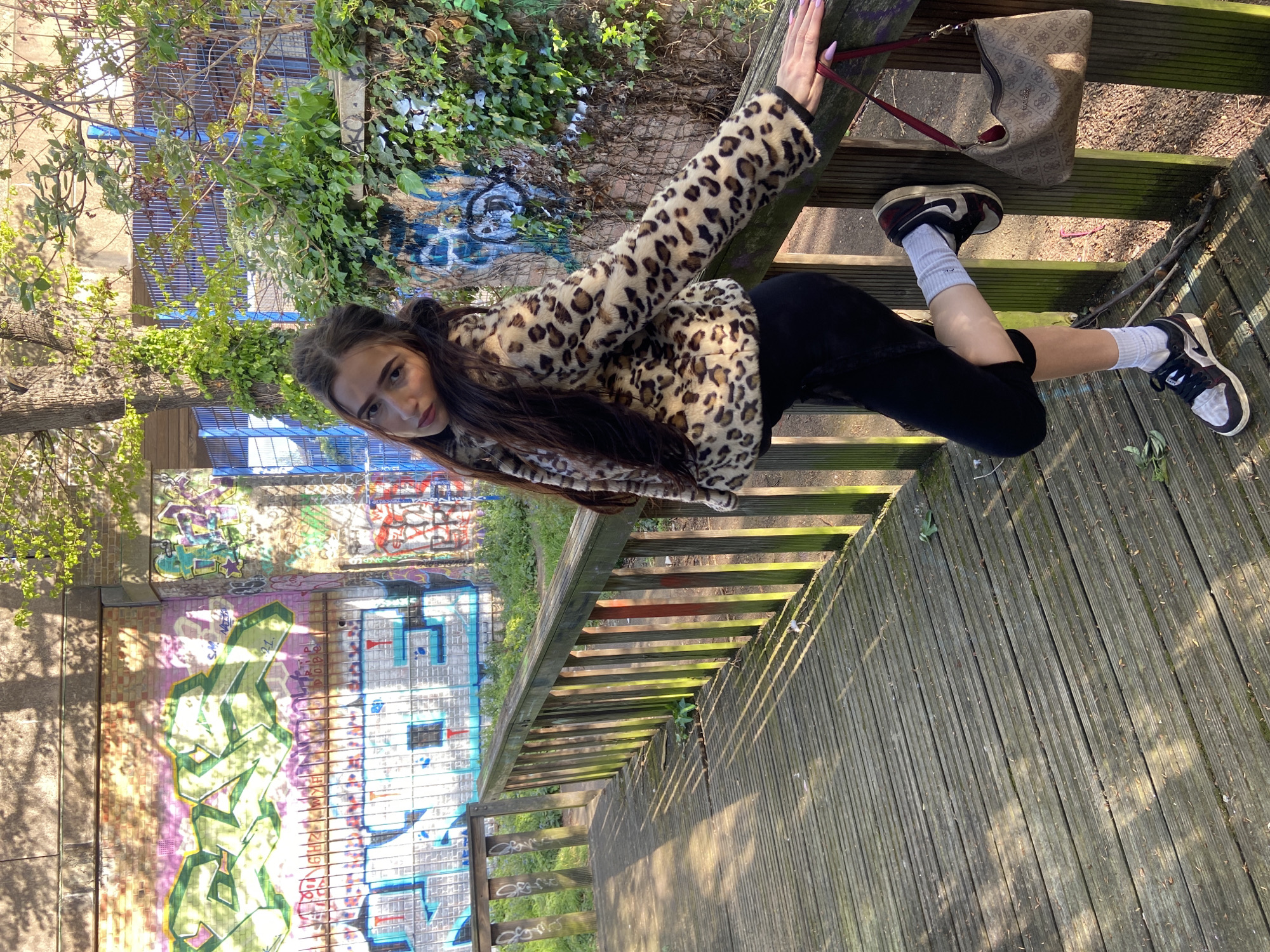 A young woman in a leopard print jacket and black leggings poses on a wooden walkway in what appears to be a park or urban area. She is leaning against the railing of the walkway, which has a wooden plank construction. Her left arm is extended, and her right leg is bent. She is wearing socks and sneakers. A brown purse is resting on the steps of the walkway. In the background, there is a wall covered in graffiti, indicating an urban setting. There are also trees and greenery visible, suggesting a park or green space within the city. Given the presence of graffiti, this image might be taken in an area of London where street art is prevalent, such as Shoreditch or Camden.