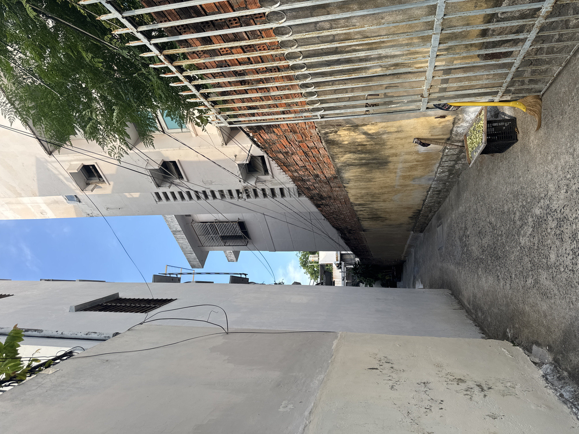 The image depicts a narrow alleyway between two buildings in Nha Trang Ward, Vietnam. The left side of the alley is a rough, off-white concrete wall with dark stains and some patches of peeling paint near the ground. Electrical wires are visible running vertically along this wall and overhead. The right side of the alley is bordered by a weathered, yellow-beige painted wall topped with a brick fence, which has a rusty metal gate on the right. Leaning against the gate is a yellow-handled broom. In front of the gate and to the left of it, there is a black crate filled with green leaves, and a wooden tray holding more green leaves rests on top of the crate. Another broom with a dark handle leans against the yellow wall behind the crate. Beyond the yellow wall, a tall, cream-colored building with multiple windows and a row of vents is visible, along with greenery from a tree. The sky above is clear and blue. The ground of the alley is paved with textured concrete.