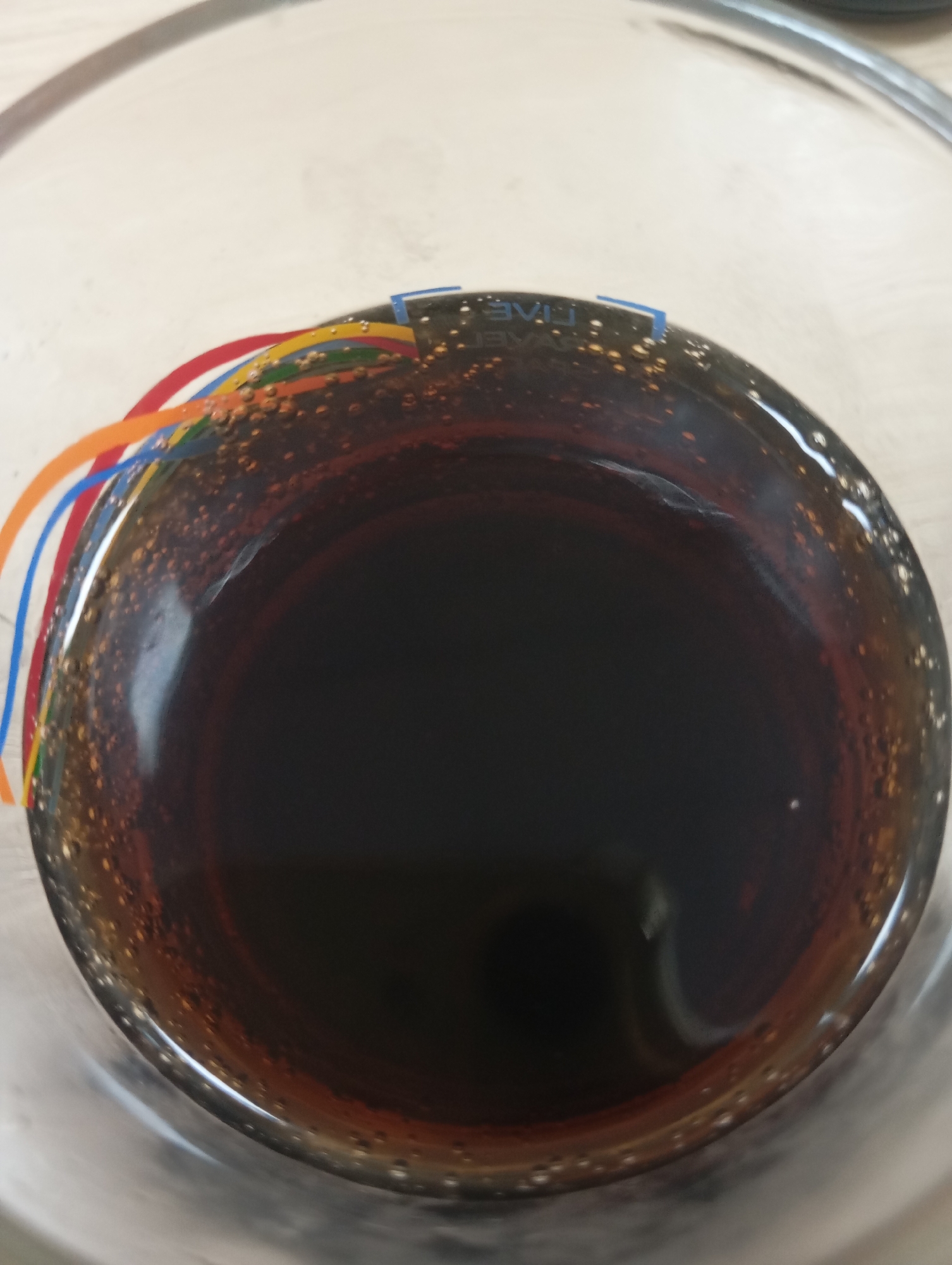 The image shows a close-up, overhead view of a glass containing a dark liquid, possibly coffee or soda, with small bubbles adhering to the inner surface. Several colored wires, red, orange, yellow, green, blue, and brown, are entering the liquid from the left side of the frame. At the top edge of the glass, a small piece of electronic component with blue and white markings is visible. The background is a light-colored, textured surface, suggestive of a table or countertop. There are no people or identifiable landmarks of Almaty, Kazakhstan visible in the image.