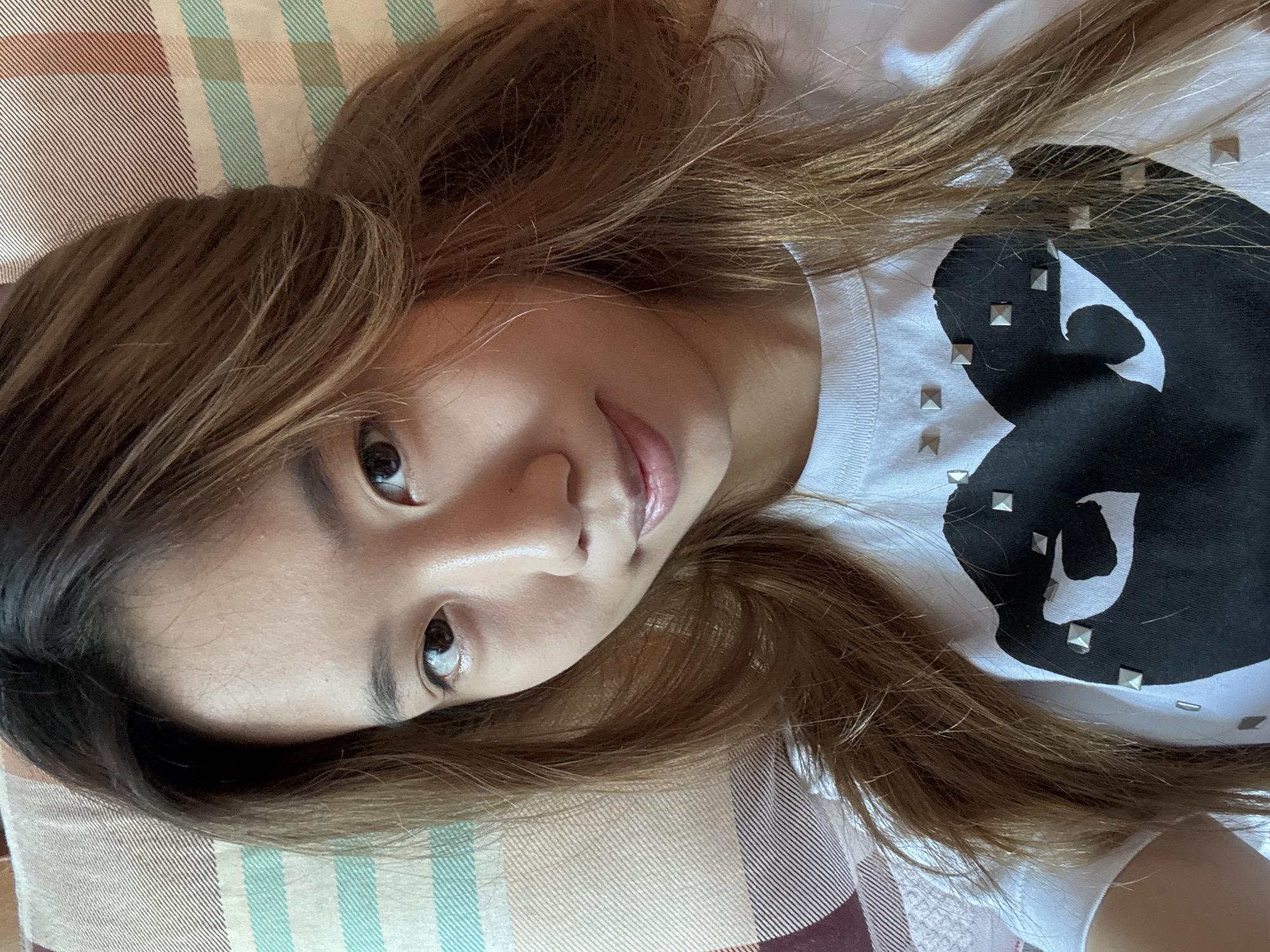 This media file is a close-up, overhead shot of a young woman lying on a striped surface. She has long, light brown hair that falls around her face and shoulders, partially obscuring her features. She is looking towards the camera with a soft, slightly amused expression. Her eyes are dark and appear to be looking directly at the viewer. She is wearing a white t-shirt with a large, black graphic design on it, possibly a stylized heart, embellished with small, square silver studs.
The background surface appears to be fabric, with a pattern of alternating stripes in various earthy tones, including brown, beige, and pale green. The lighting suggests it could be daytime, possibly indoors, given the soft, diffused light. The overall mood is relaxed and intimate, with the woman appearing comfortable and at ease. There is no visible text in the image. The location context provided is Guandu, China, though there are no specific visual cues within the image to confirm this.