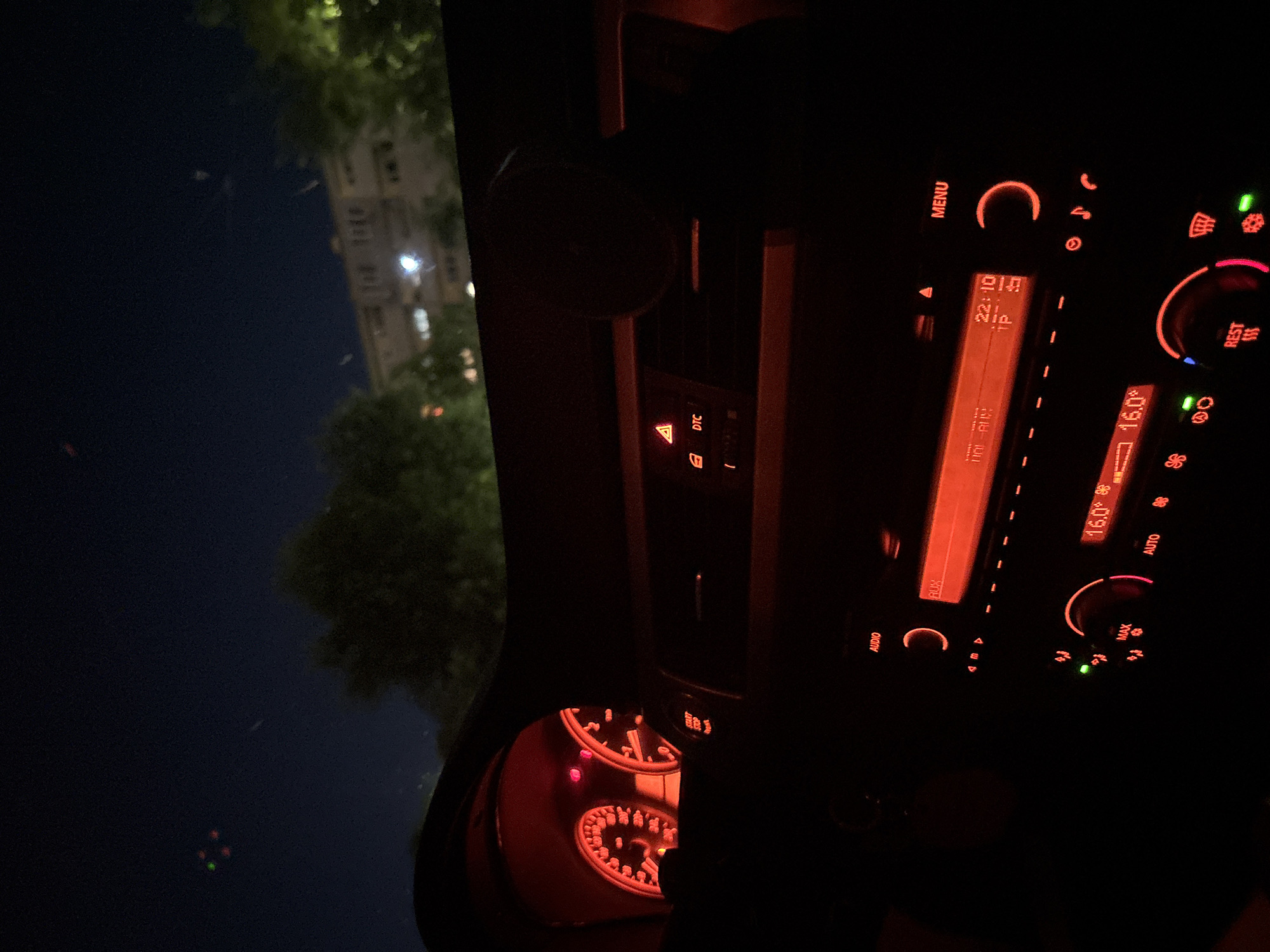 The image shows the interior of a car at night. The focus is on the dashboard and center console, which are illuminated by a warm orange glow from the instrument panel. The time displayed on the car's infotainment system is 22:10, and the temperature is 16.0 degrees Celsius. Various buttons and controls are visible, including "MENU," "AUDIO," "AUX," "MAX," "AUTO," and "REST." There are also symbols for climate control, such as a fan and snowflake, and a hazard light button. To the left of the dashboard, a blurred view of trees and a building can be seen through the car window, suggesting an urban or suburban setting. The overall impression is one of a person inside a parked car at night, possibly taking a break or waiting.