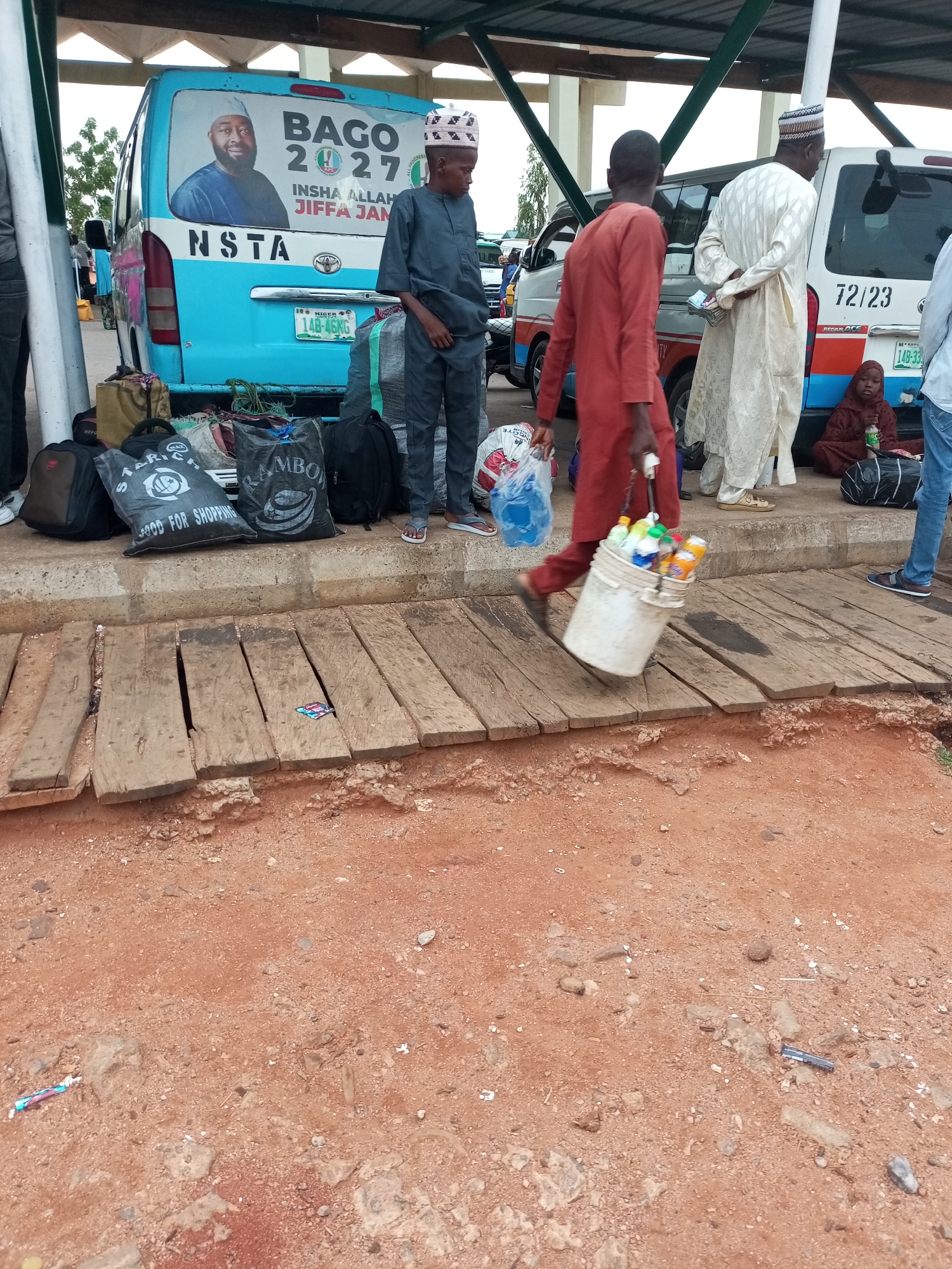 In Gombe, Nigeria, a scene unfolds at what appears to be a bus station or transportation hub. A blue and white bus, likely a commercial vehicle, is prominently featured with a large political campaign poster of "BAGO 2027" and a portrait of a man. The bus has a license plate reading "14B-6KRG". Several people are present, some appearing to be travelers with luggage, while others may be vendors. One individual, dressed in a reddish-brown traditional outfit, is walking and carrying a white bucket filled with various bottles of beverages. Another person, in a light-colored traditional outfit, stands near a white vehicle with the number "72/23" on its side. A child in a maroon hijab is seated on the ground near the vehicle. Bags of various sizes and colors are scattered on the ground, some with labels like "STARICH" and "RAMBO". The environment is characterized by rough, reddish-brown soil and a wooden walkway composed of weathered planks. The overhead structure suggests a covered waiting area. The overall impression is one of activity and transit within a busy public space.